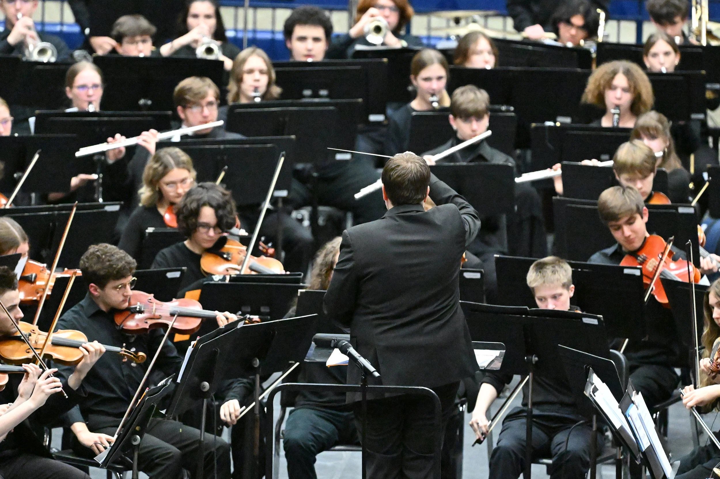 A conductor leading an orchestra of young musicians playing violins and other string instruments on stage.