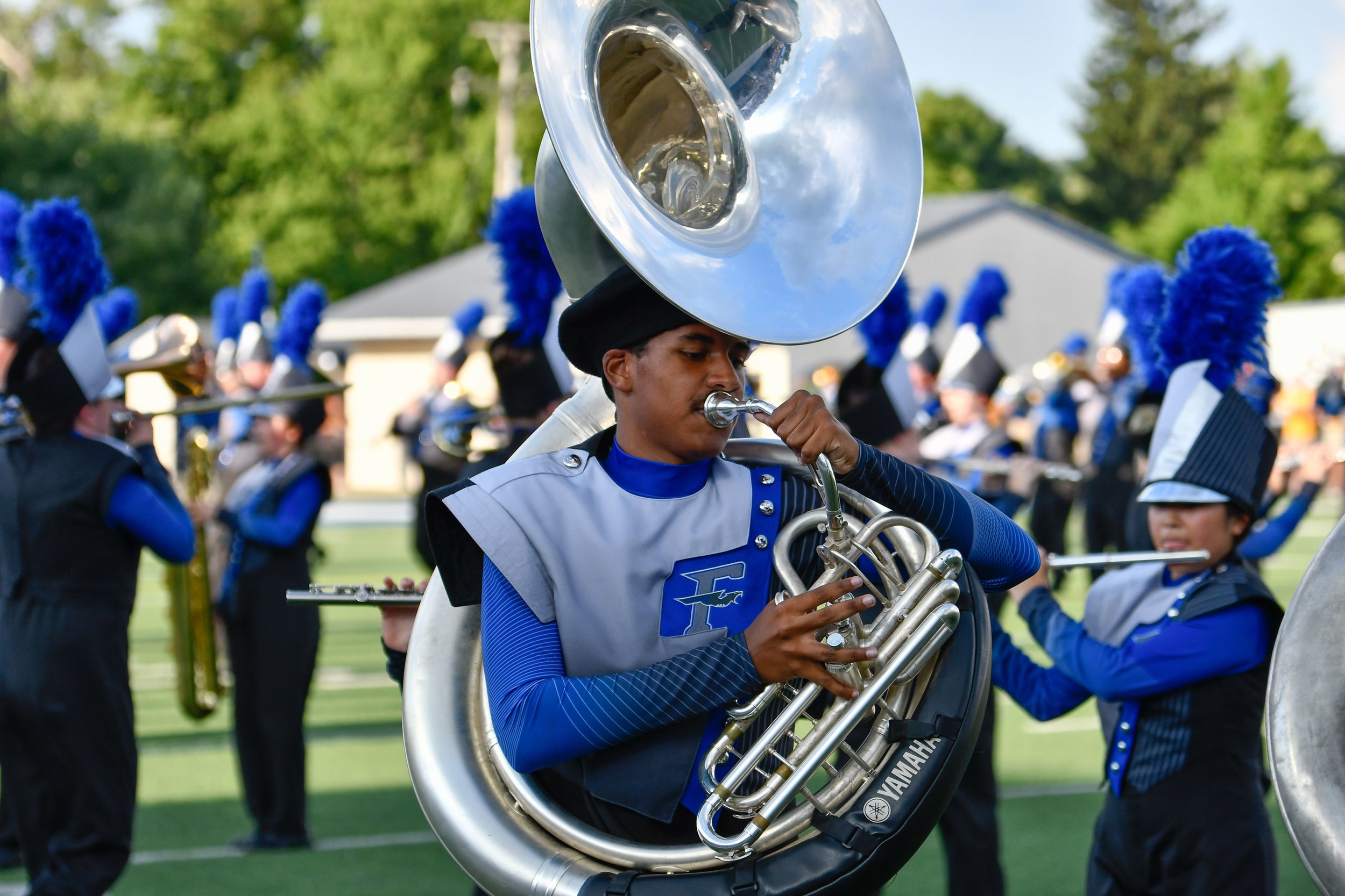 A marching band performs outdoors on a field with green trees in the background. The focus is on a young male band member playing a sousaphone, wearing a blue and gray uniform with a logo and a black hat with a blue feather.