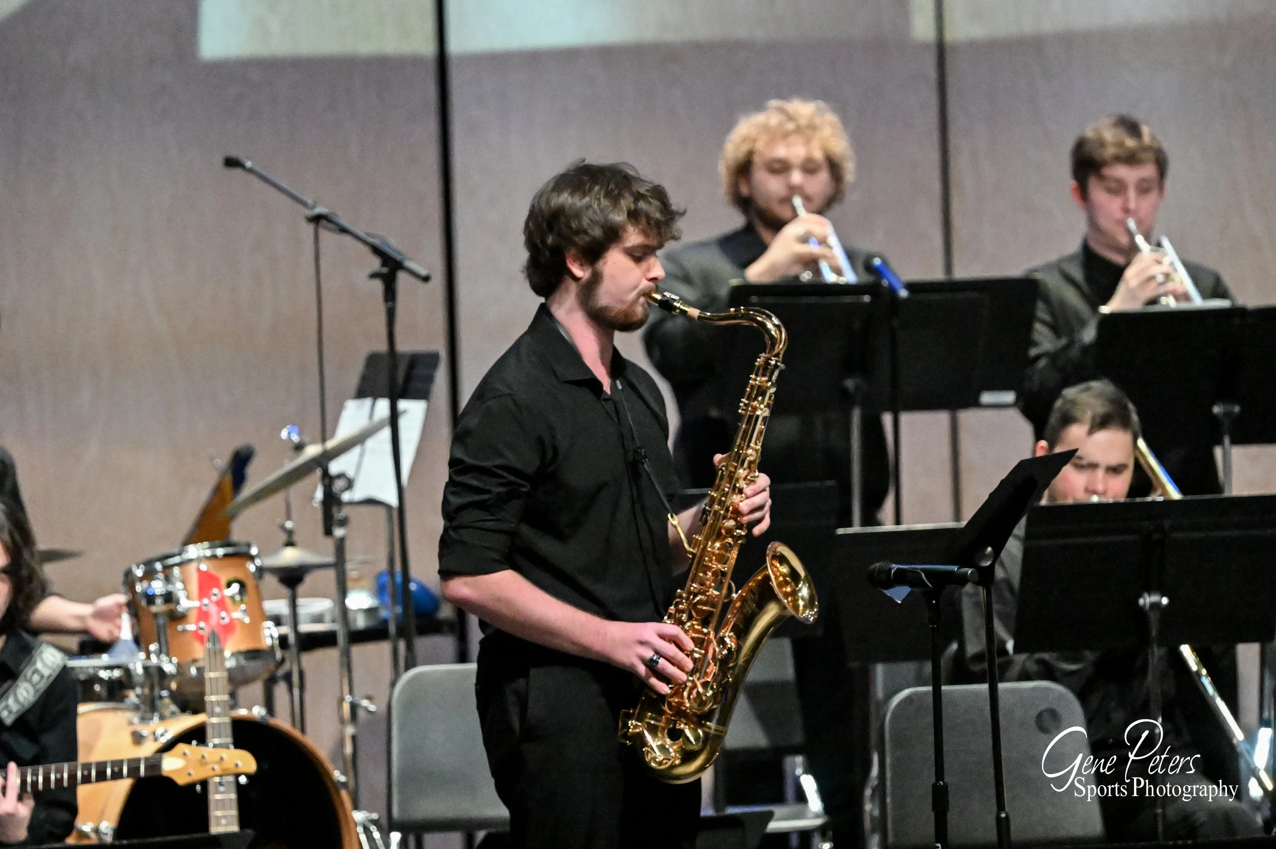 Musicians performing on stage, including a man playing an alto saxophone in the foreground and two men playing trumpets in the background.