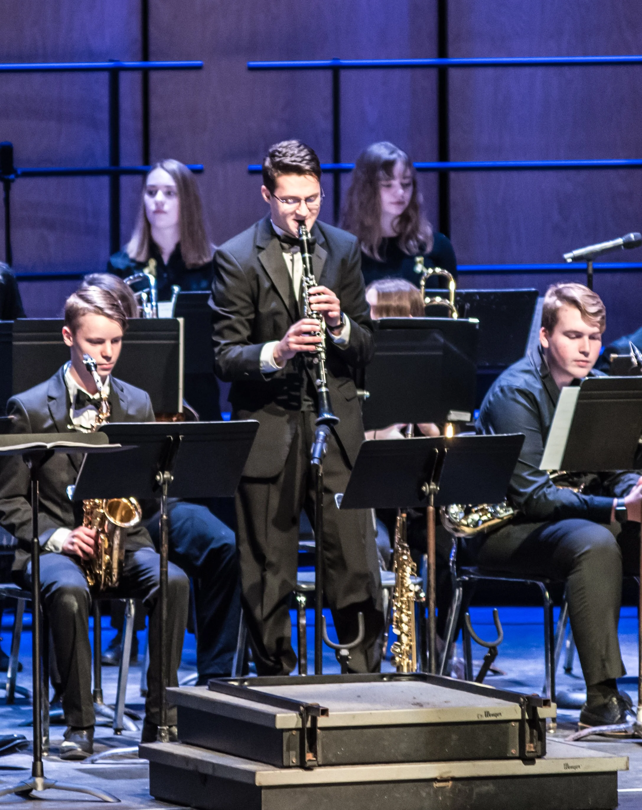 A group of young musicians performing in a concert, with a male saxophone player standing and playing while others seated with music stands around him.