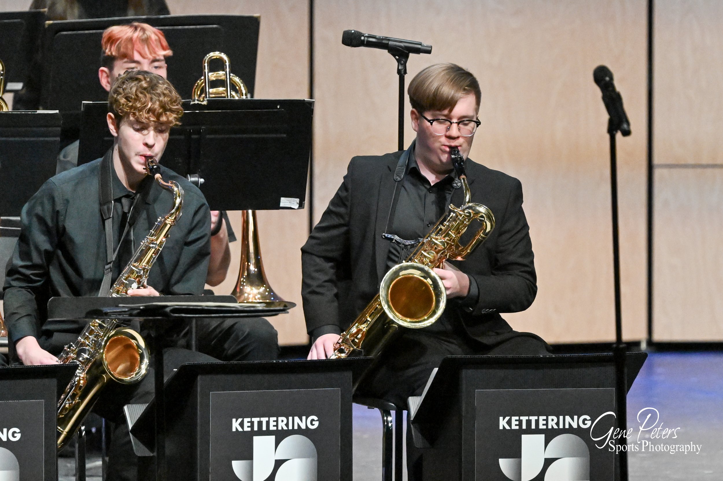 Two young men playing jazz saxophones on stage during a concert, with music stands labeled 'Kettering' in front of them, and other musicians visible in the background.