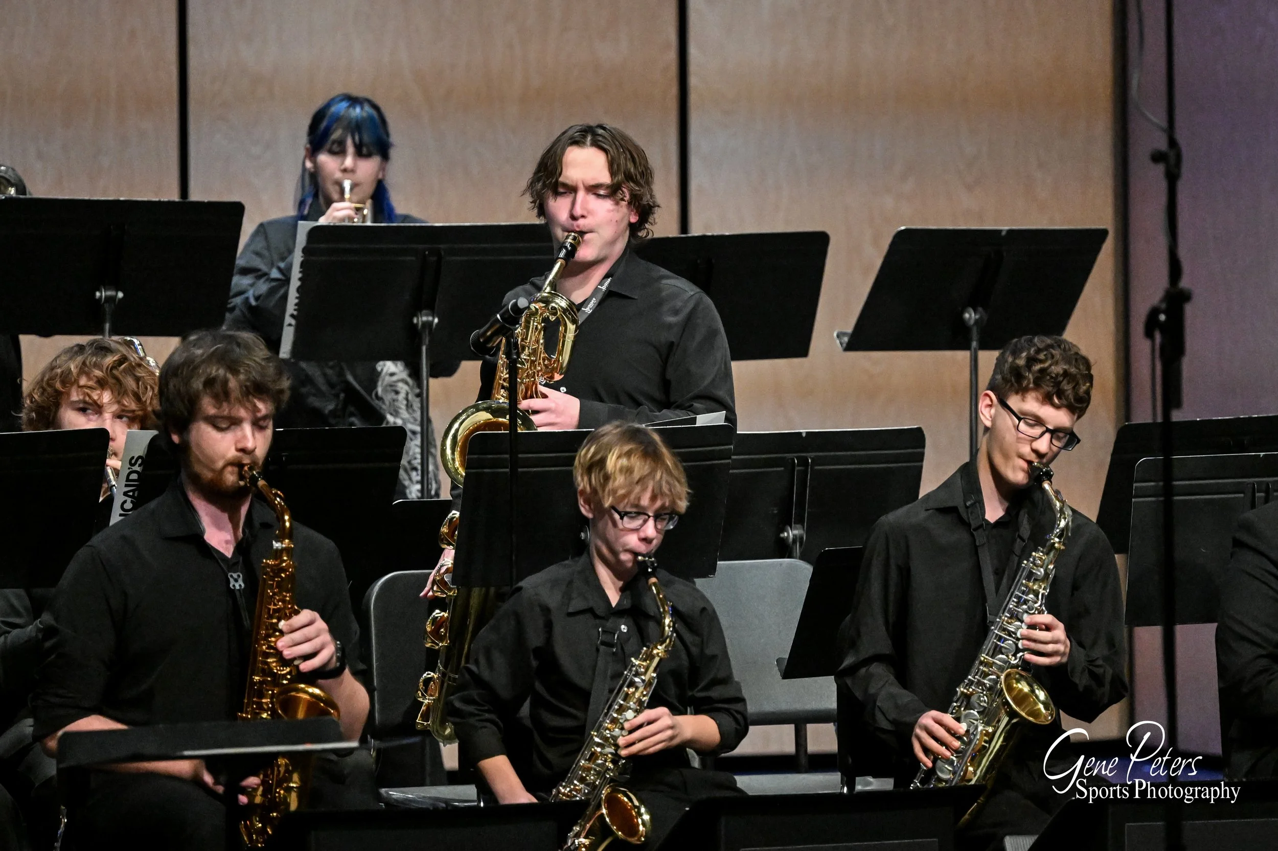 Young musicians playing saxophones on stage, with music stands and a wooden background.