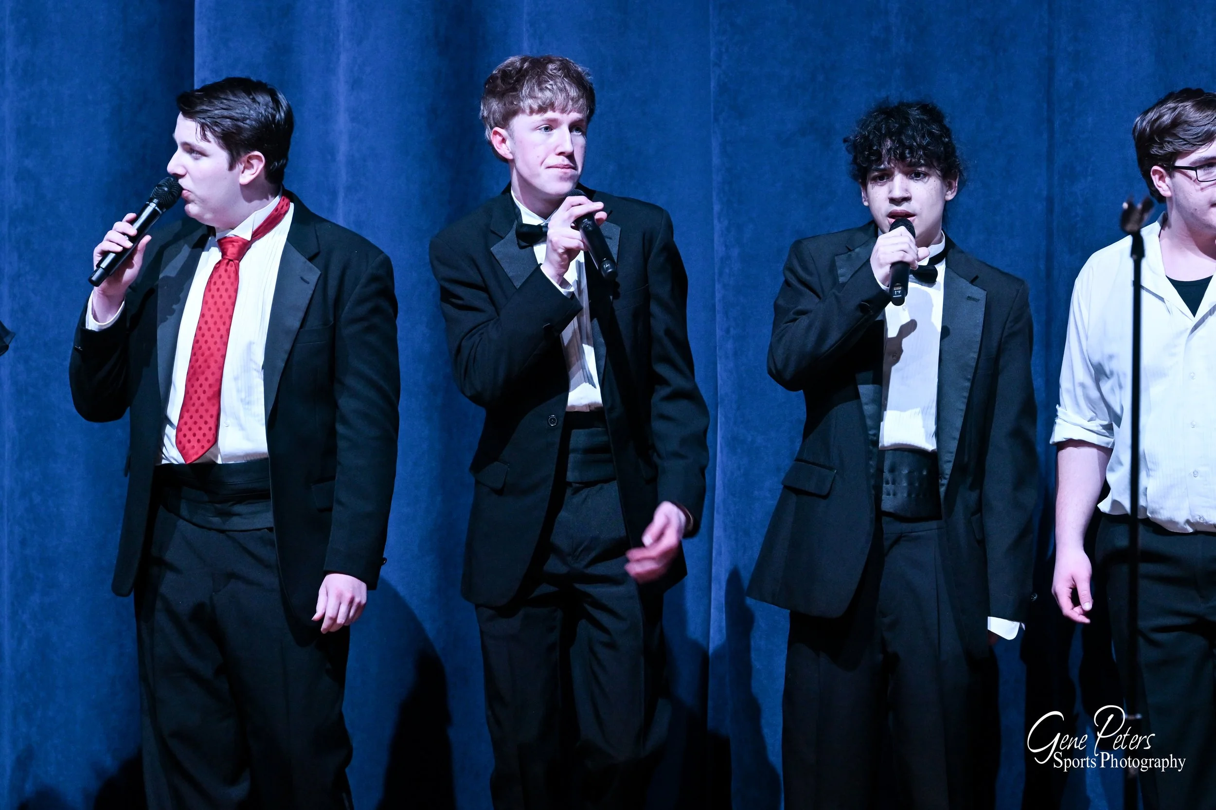 Four young men standing on stage holding microphones, dressed in formal black and white attire, against a blue curtain background.