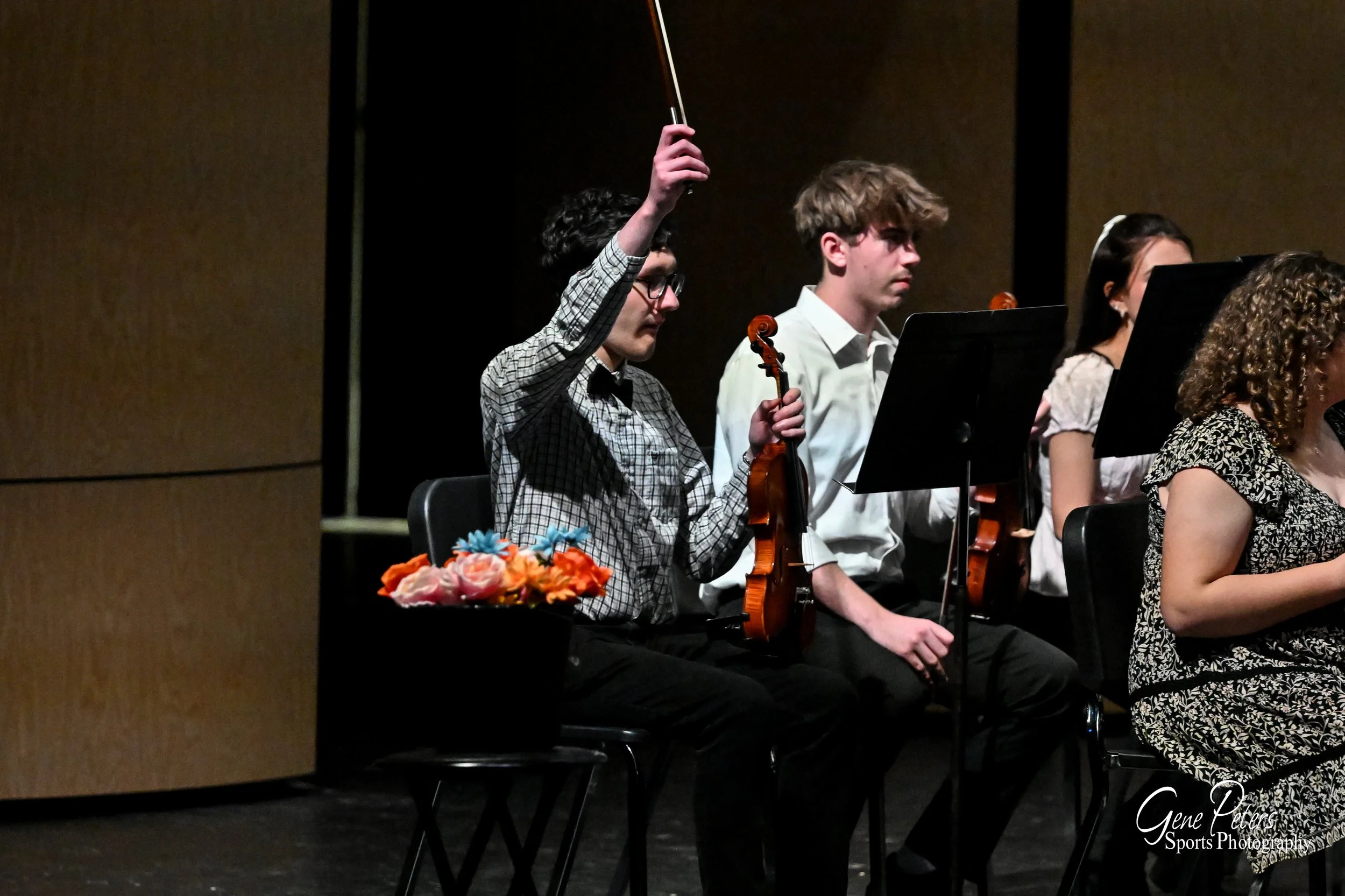 Young musicians sitting with violins, one raising a bow, during a performance or rehearsal.