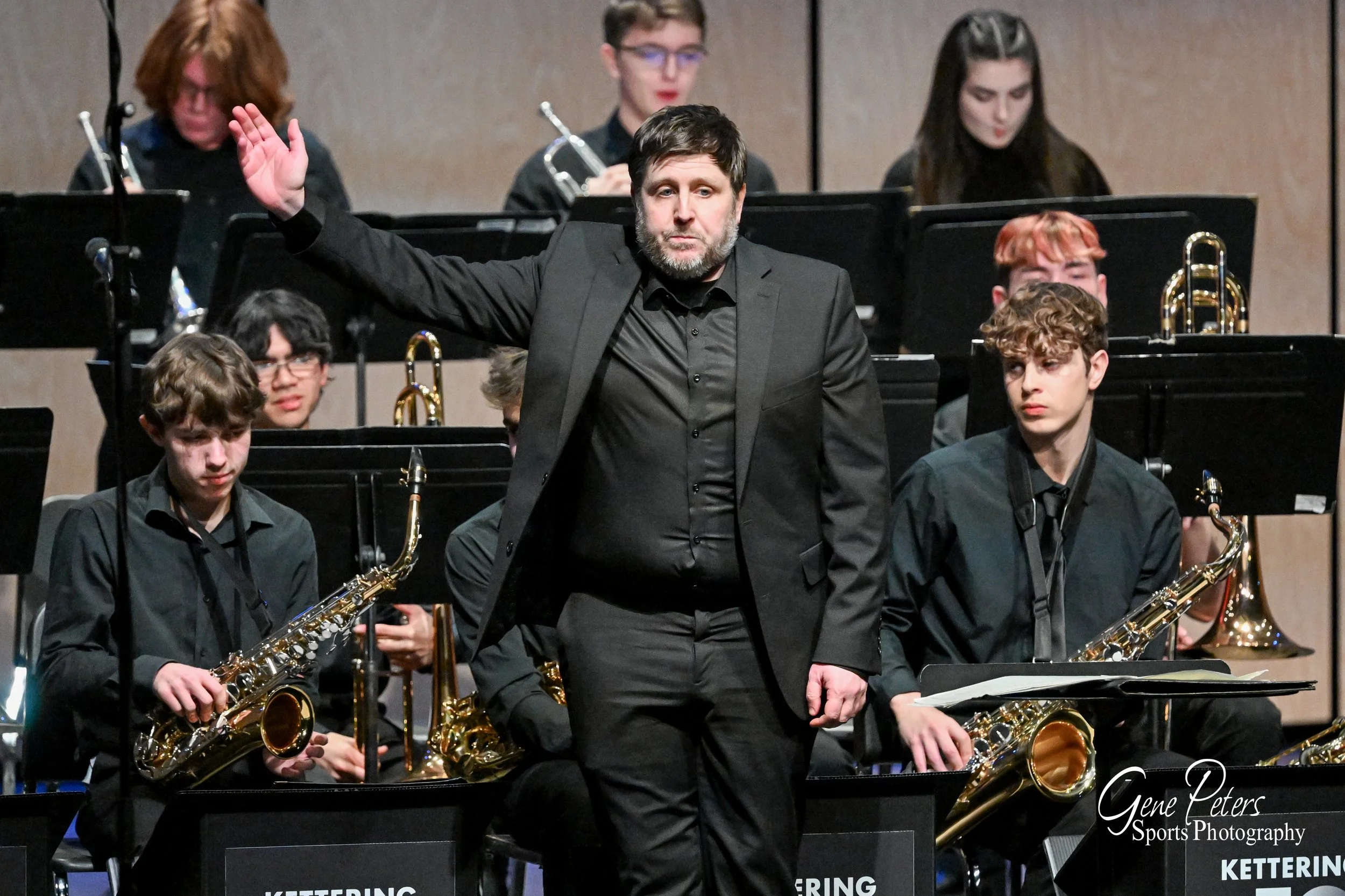 A band conductor in a black suit leading a jazz band during a performance, with young musicians playing saxophones and other instruments in the background.