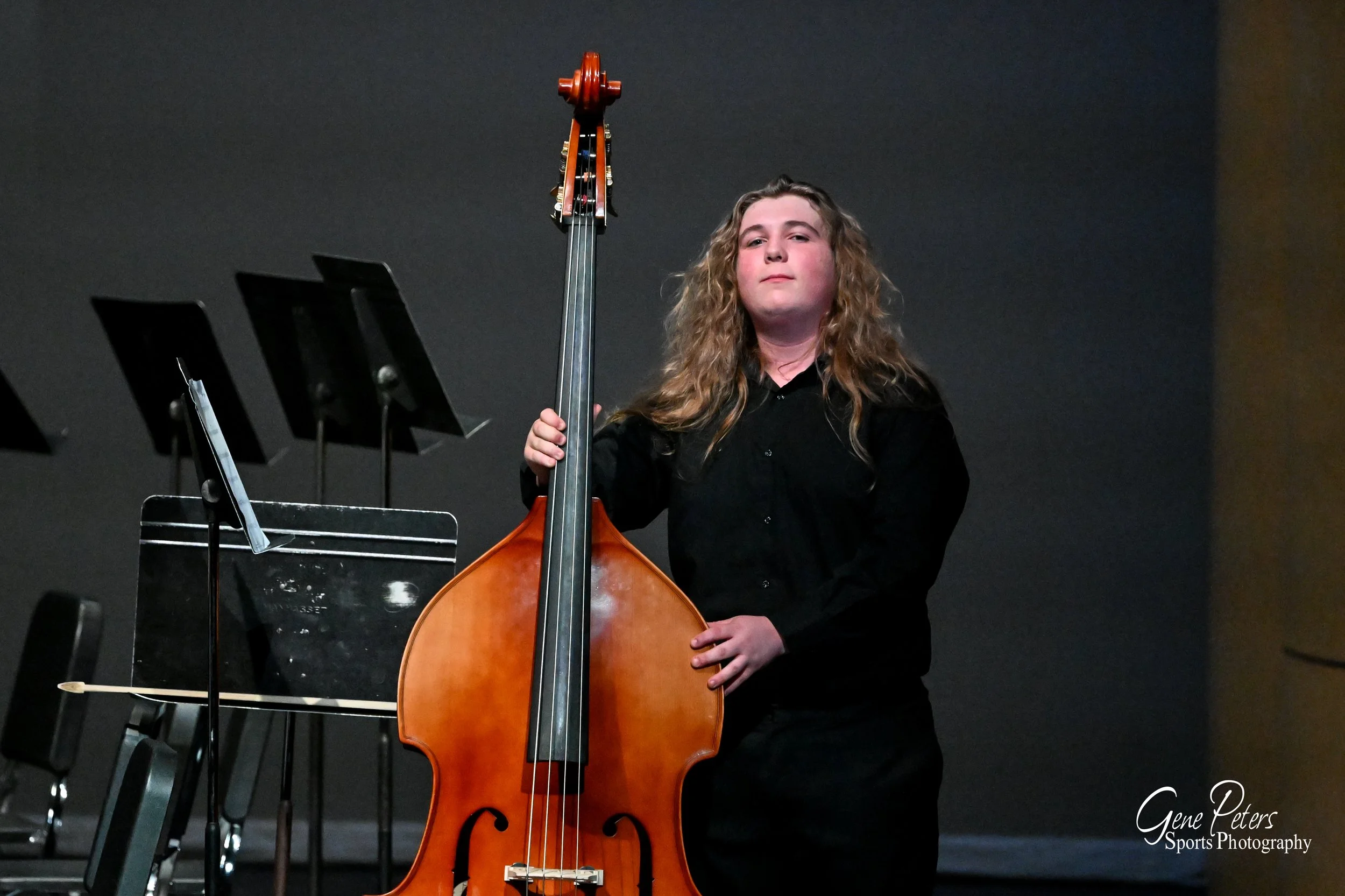 Young girl with long curly hair wearing a black shirt playing a double bass on stage at a concert or recital.