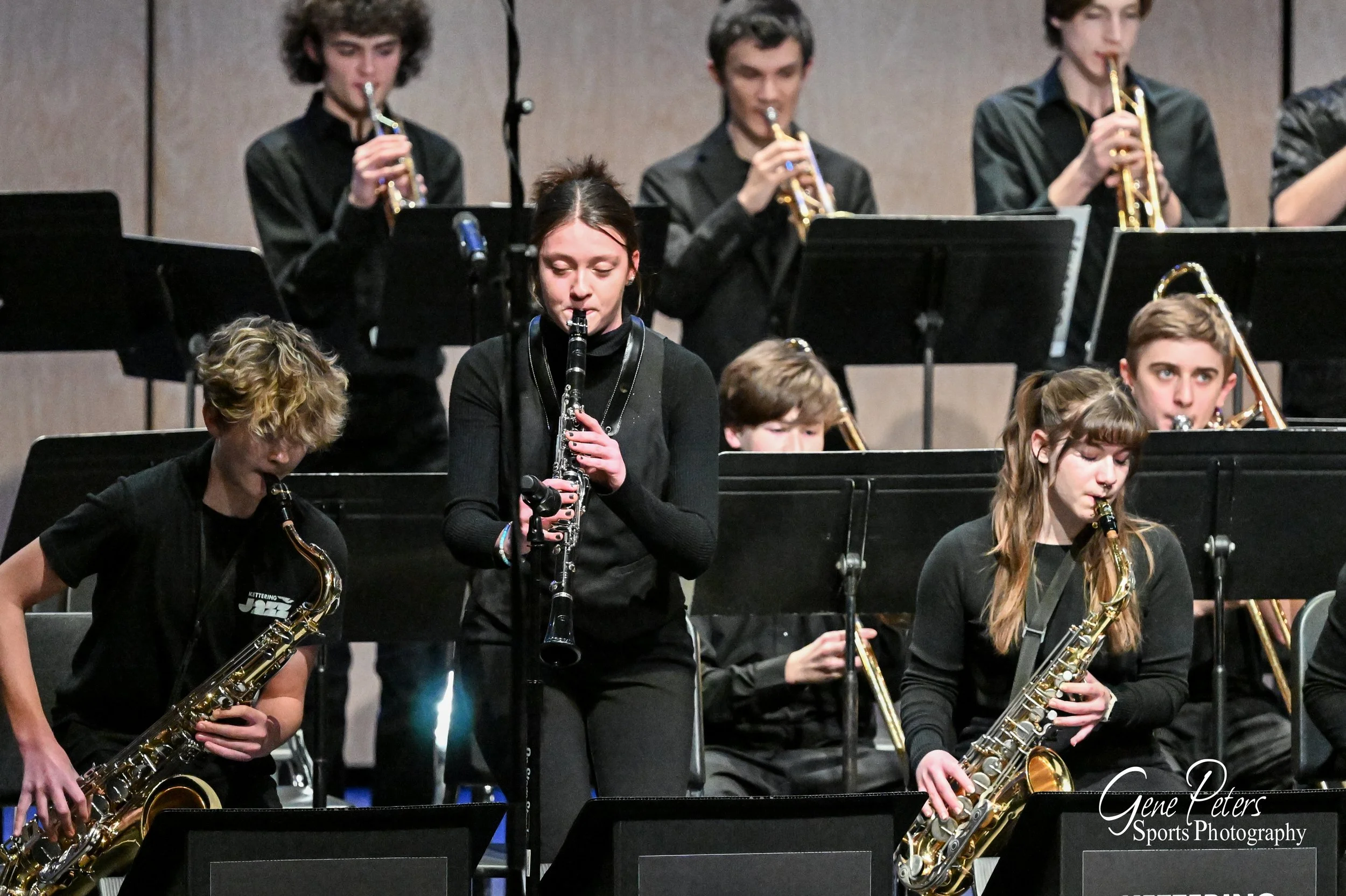 Youth orchestra performing on stage with musicians playing saxophones, trumpets, and clarinets, dressed in black, in a concert hall.
