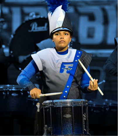 A young boy in a drum major uniform, holding drumsticks, standing in front of a drum set at a parade or marching band event.
