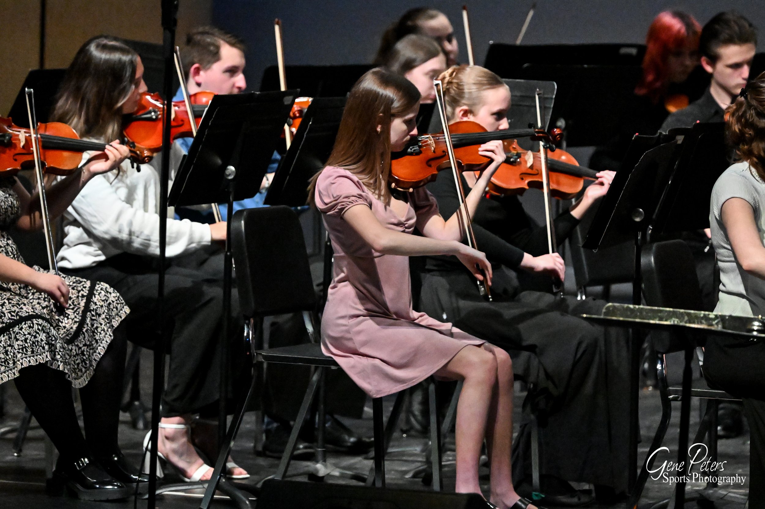Orchestra group performing on stage with violinists seated and playing their instruments.