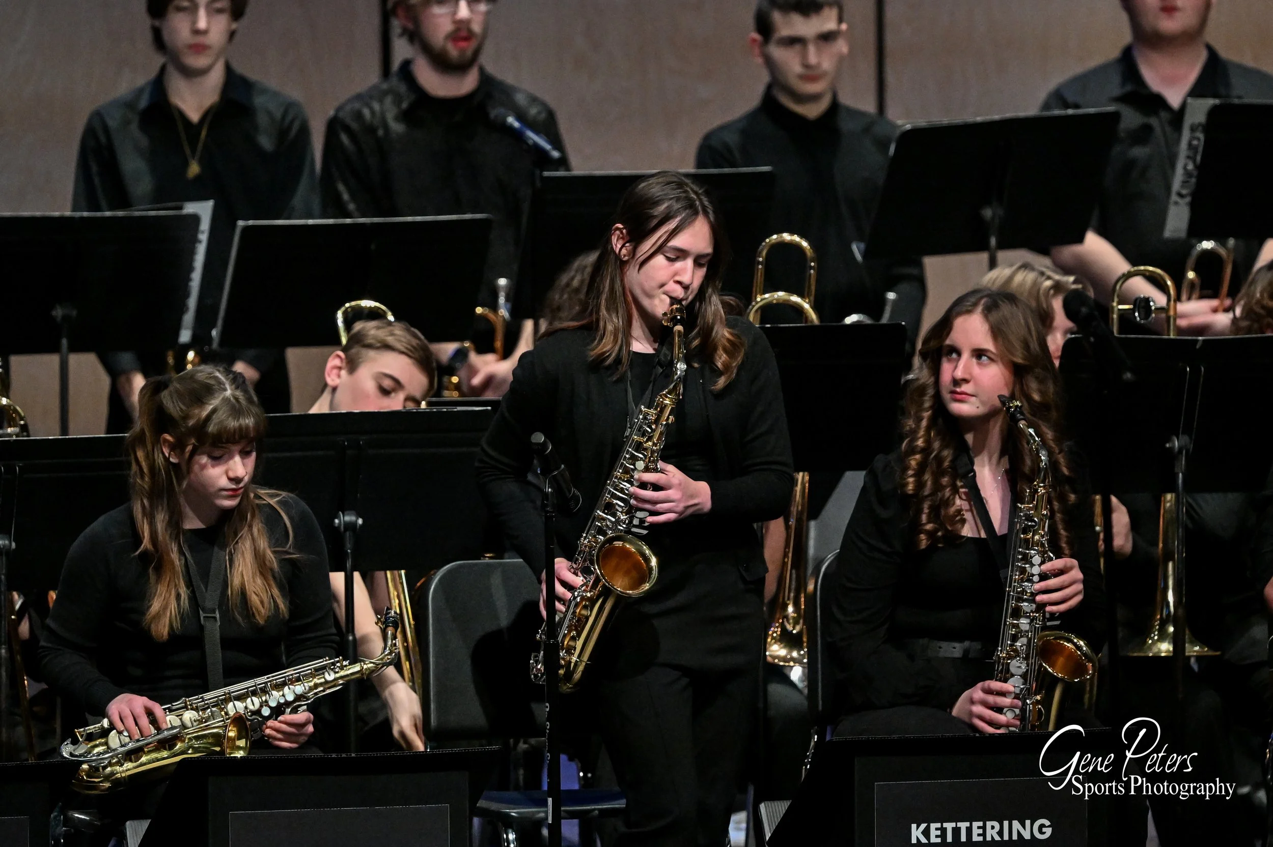 Young female musicians holding saxophones during a concert, with other musicians and sheet music stands in the background.