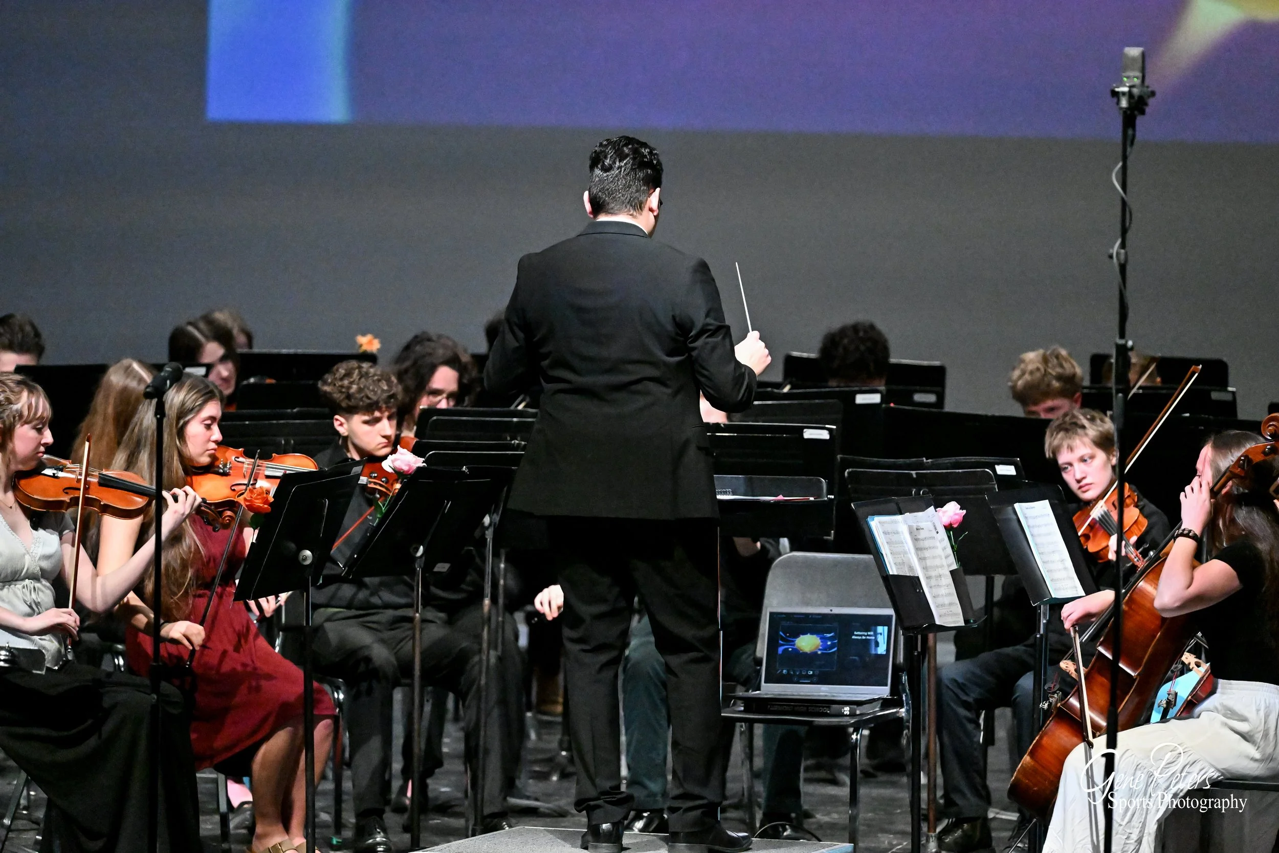 An orchestra performing on stage, led by a conductor in a black suit with a baton, with musicians playing violins, cellos, and other instruments, and a laptop on a chair in front of the conductor.