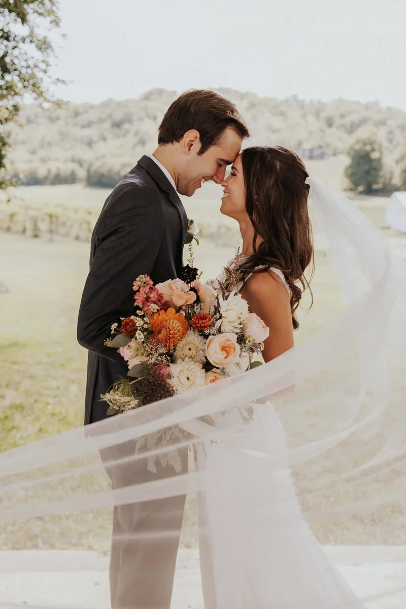 A newlywed couple is embracing outdoors, with the bride holding a large bouquet of colorful flowers, and they are touching foreheads with smiles.