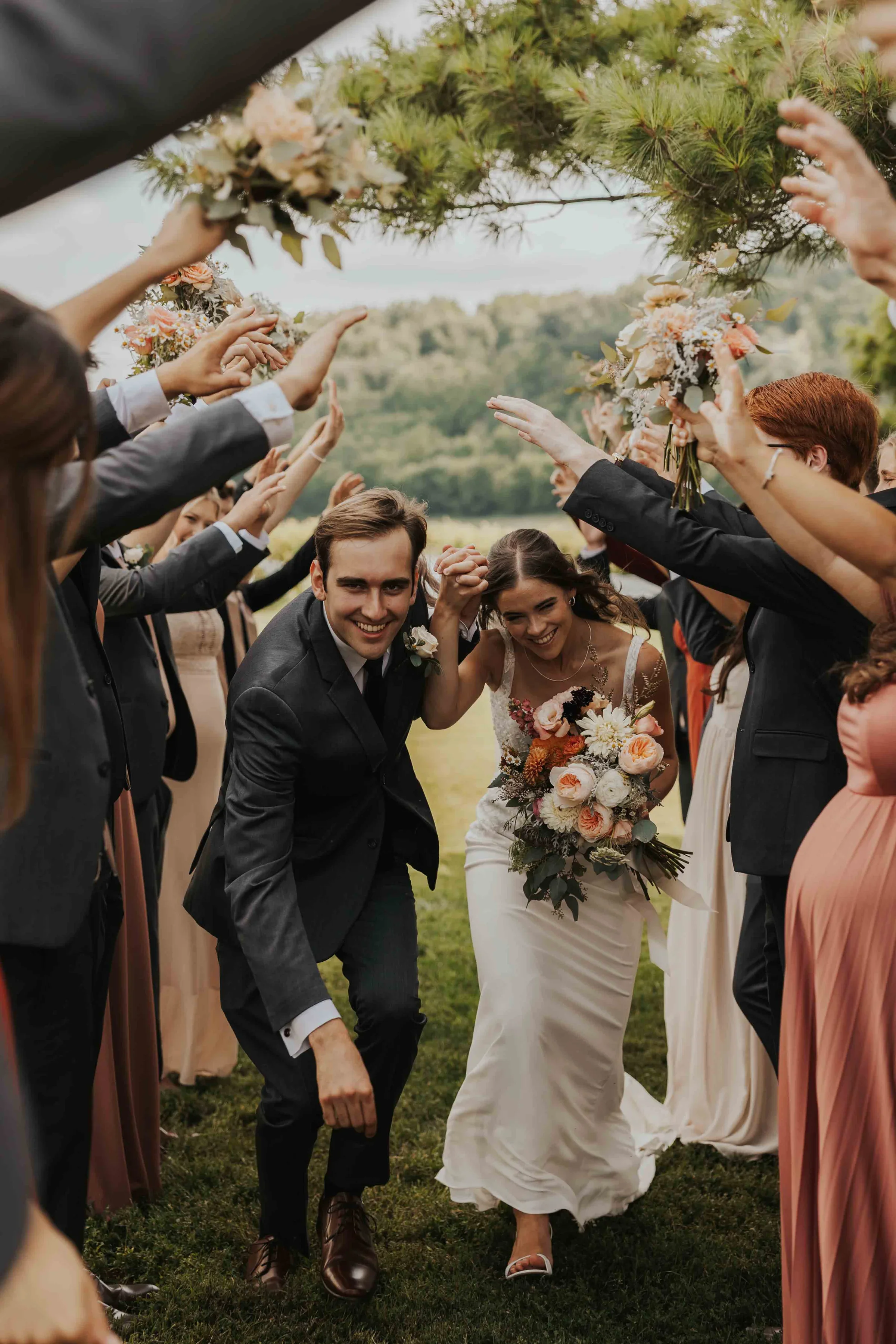 Happy newlywed couple walking between wedding guests, holding hands and smiling. The bride is dressed in a white gown and carrying a bouquet, while the groom is in a dark suit. Guests are reaching out with flowers and smiling.