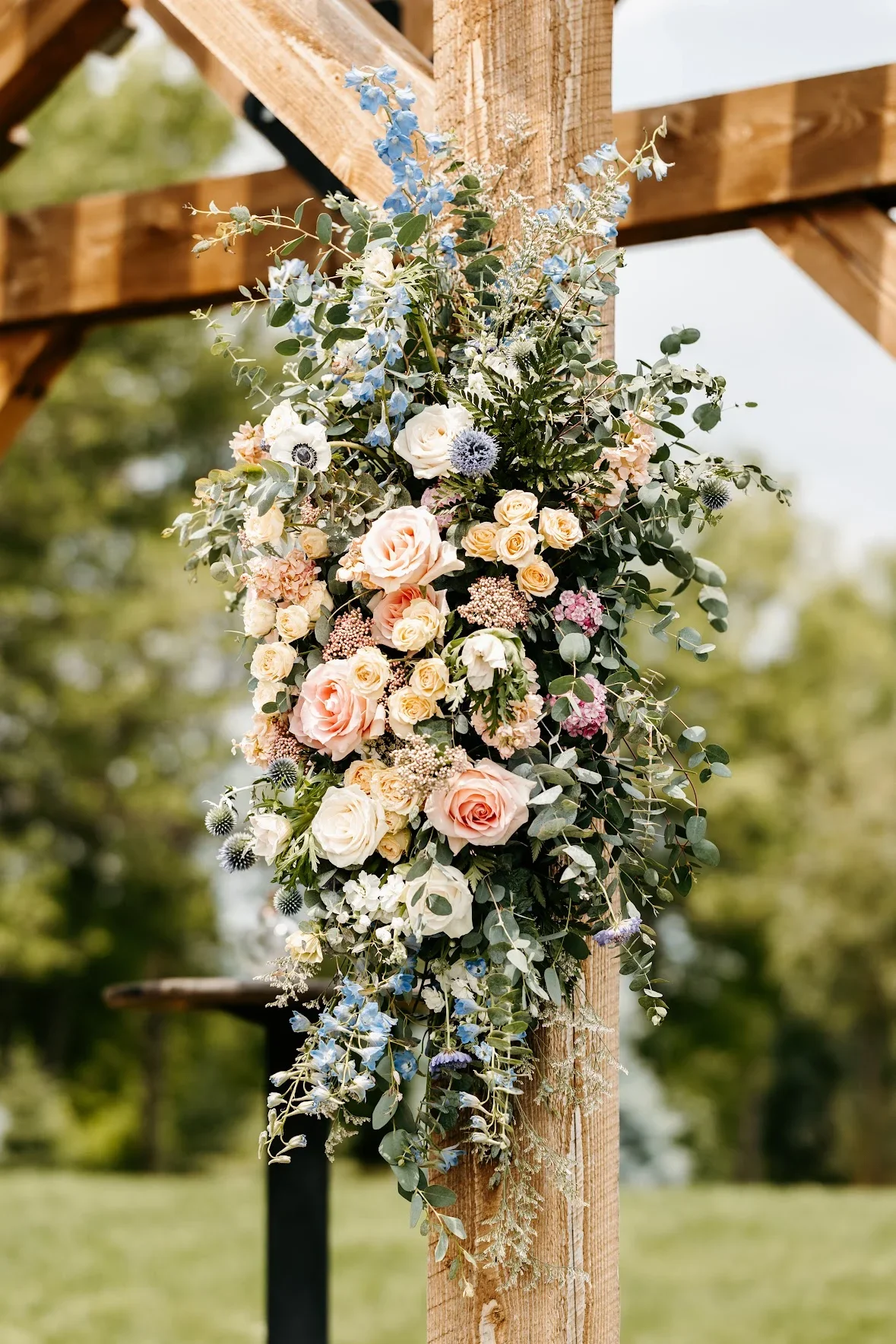 A floral arrangement hanging from a wooden structure, featuring pink and white roses, blue delphiniums, and various green foliage.