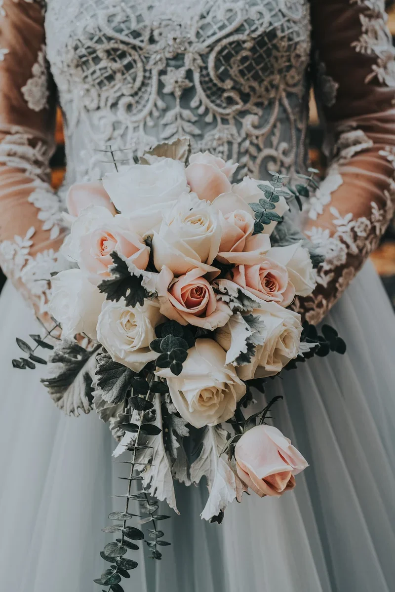 A bride holding a bouquet of white and pale pink roses with green foliage, in front of a wedding dress with intricate lace and embroidery details.