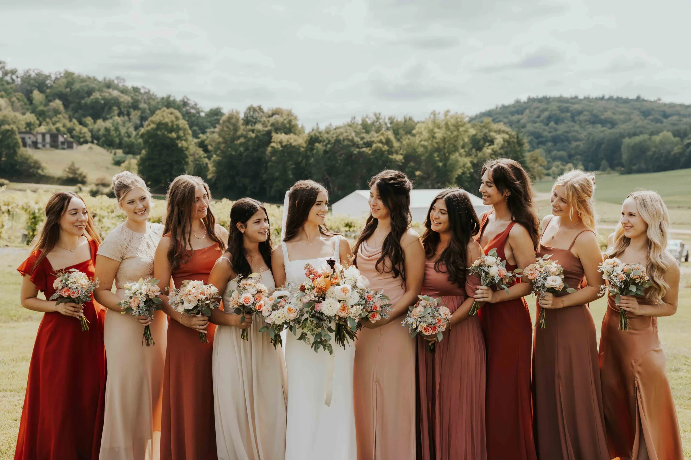A group of women, including a bride in a white dress and bridesmaids in various shades of red and pink dresses, standing outdoors on a green field with trees and hills in the background. They are holding bouquets of flowers and smiling at each other.