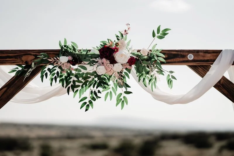 Wooden arch decorated with white drapes and a floral arrangement of roses and greenery, set outdoors.