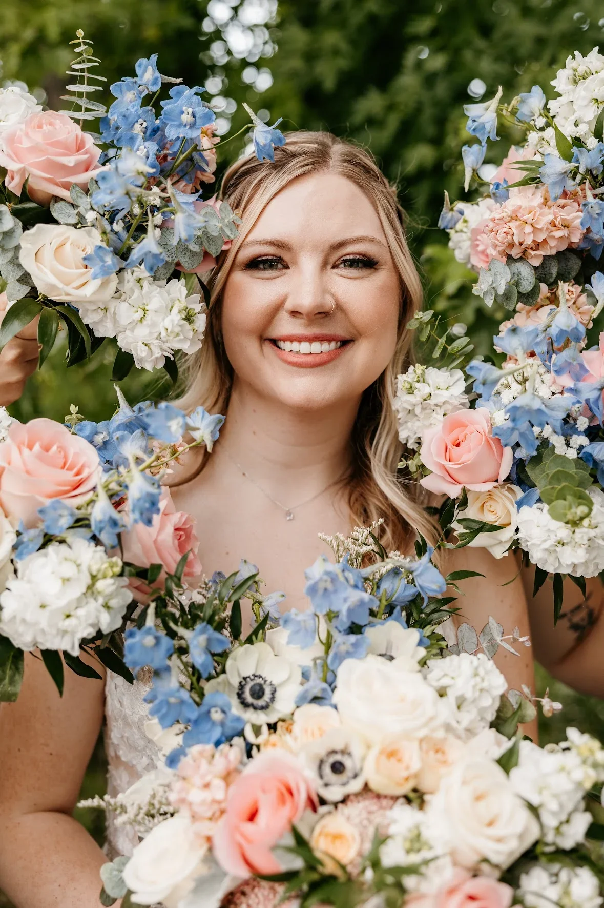 A smiling woman holding a large bouquet of pastel-colored flowers, surrounded by more flowers in a garden setting.