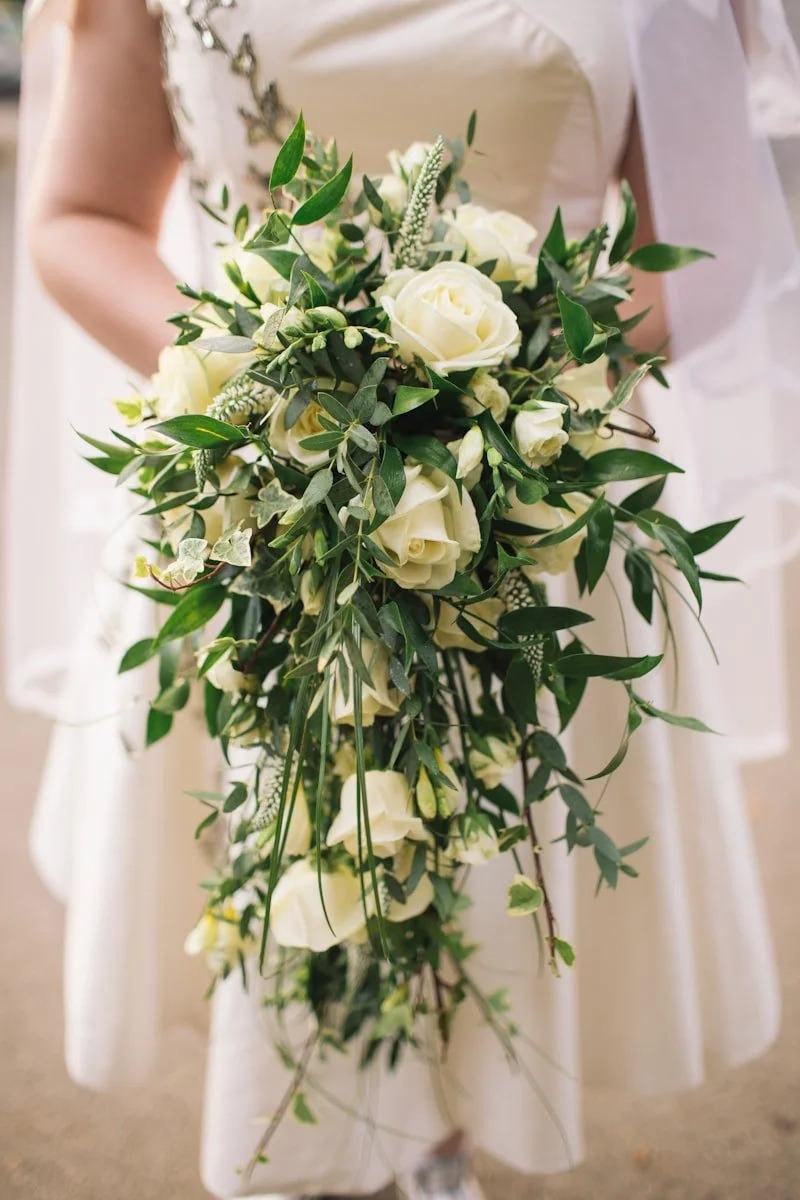 A woman holding a cascading bouquet of white roses and green foliage, with a white dress.