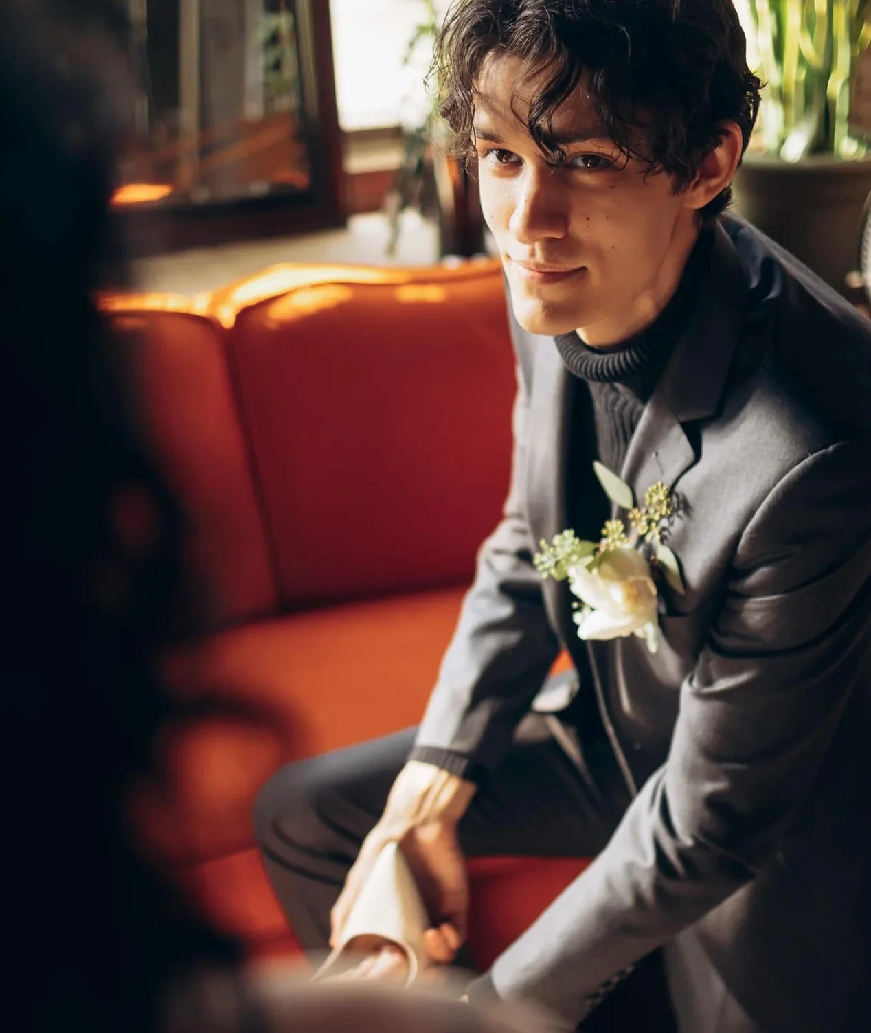 A young man in a gray suit with a white boutonniere is sitting on an orange couch, smiling softly at someone off-camera.