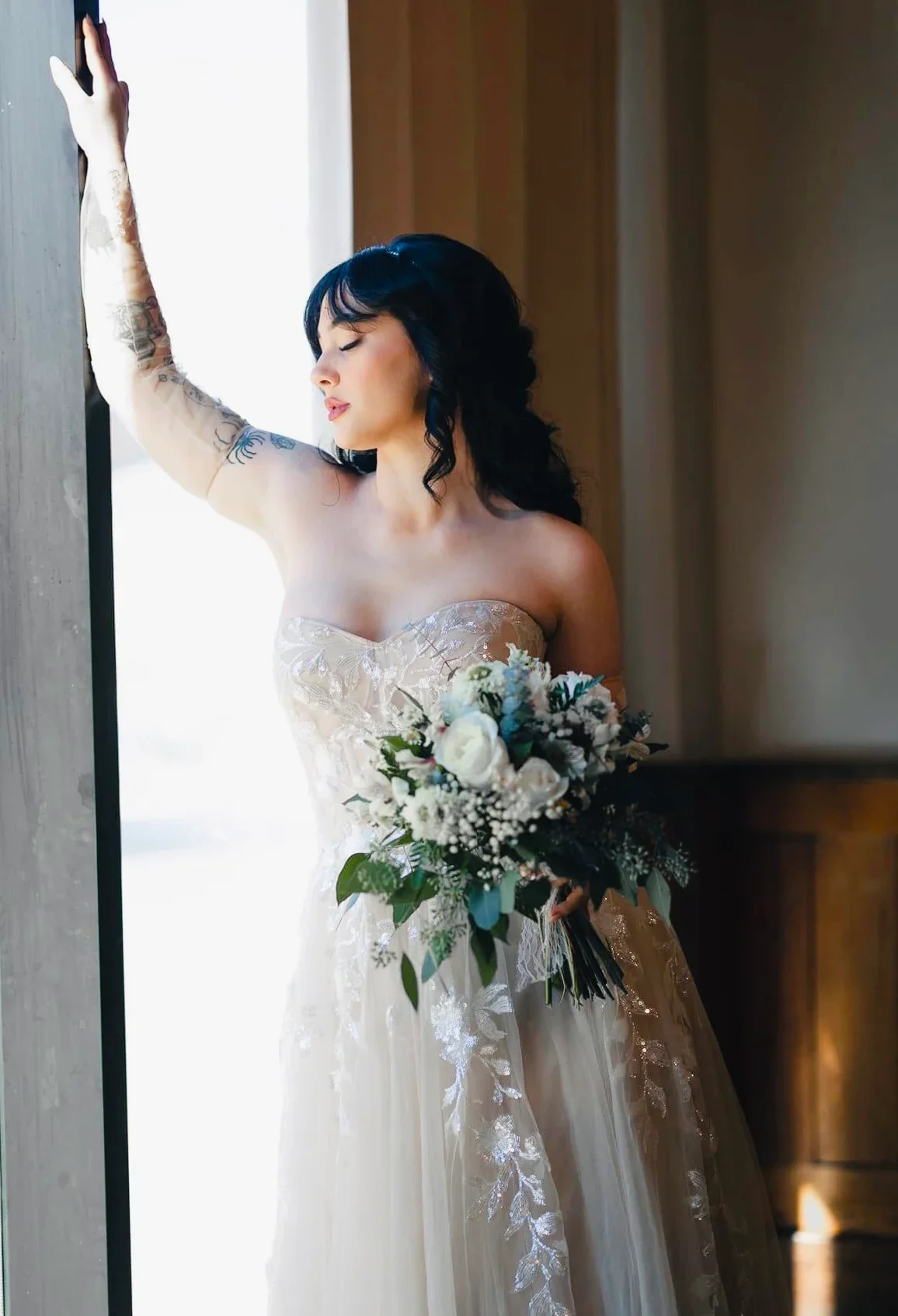 A bride in a strapless wedding dress with lace details, holding a bouquet of white flowers, standing by a window with her eyes closed and one arm raised touching the window frame.