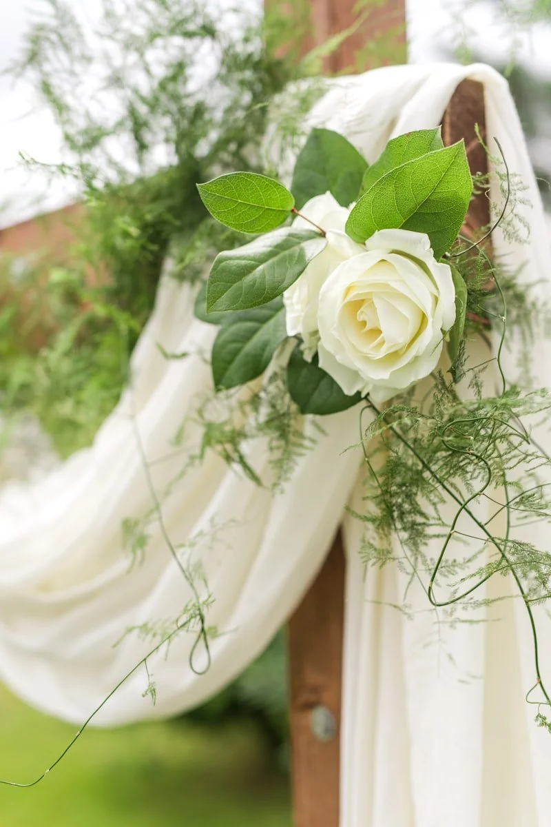 A white rose attached to a wooden post, decorated with green leaves and flowing white fabric surrounded by greenery.