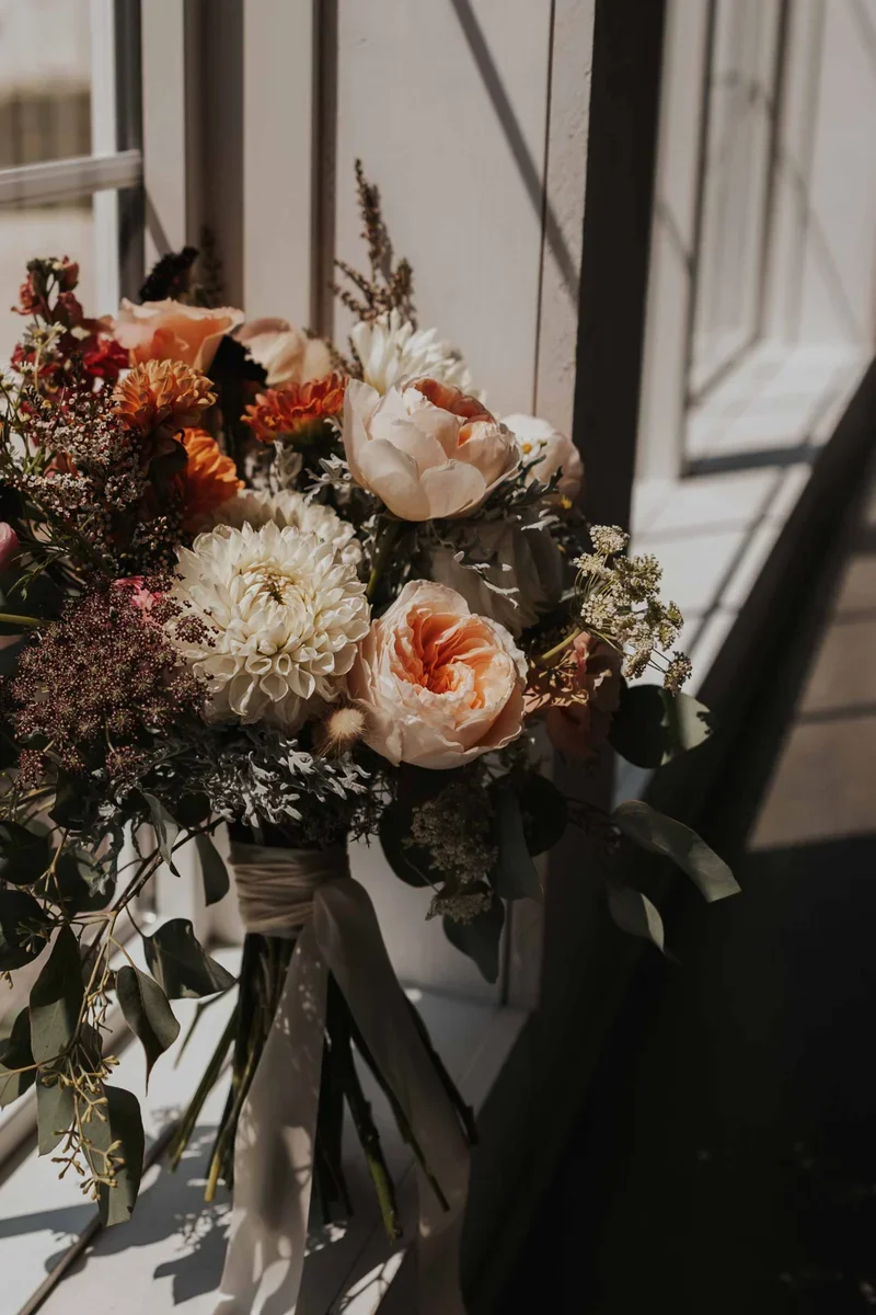 A bouquet of mixed flowers including peonies, dahlias, and other blooms, tied with a ribbon, placed near a window with sunlight casting shadows.