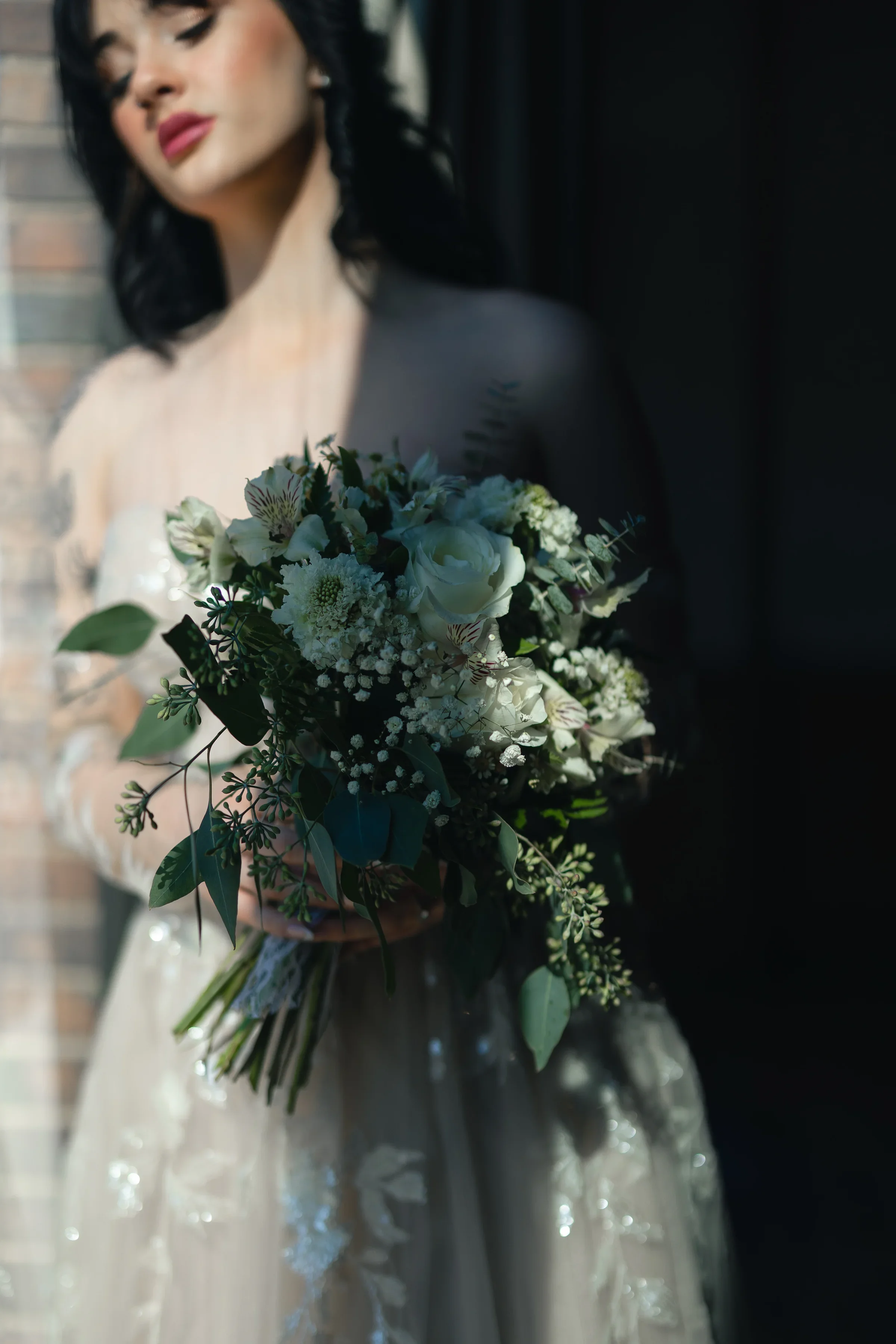 A woman in a wedding dress holding a bouquet of white and green flowers, standing against a brick wall with shadows.