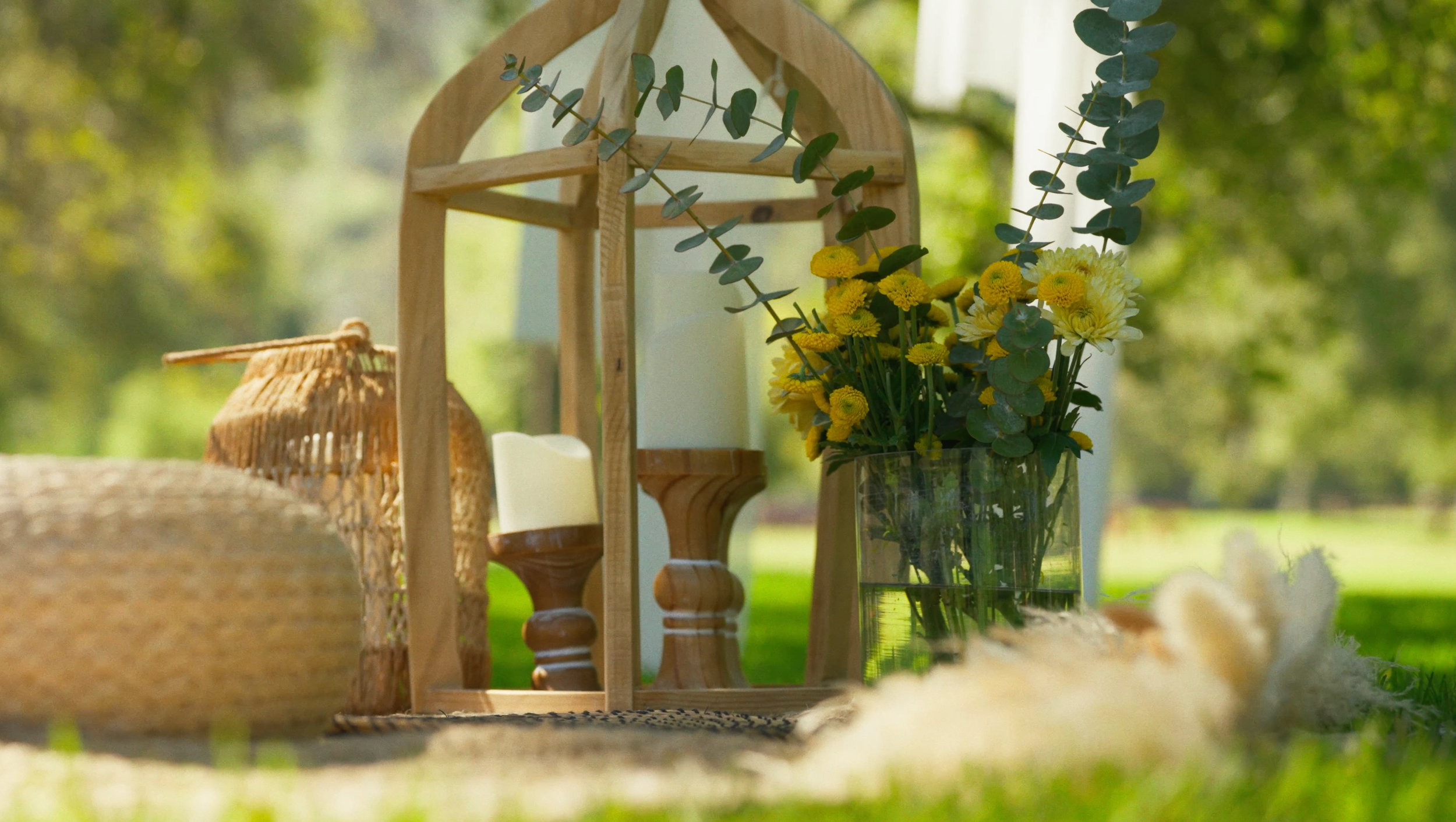 A decorative outdoor setting featuring a clear glass vase with yellow flowers and green foliage, a wooden lantern with a candle inside, and woven baskets on a table, with a blurred green garden background.