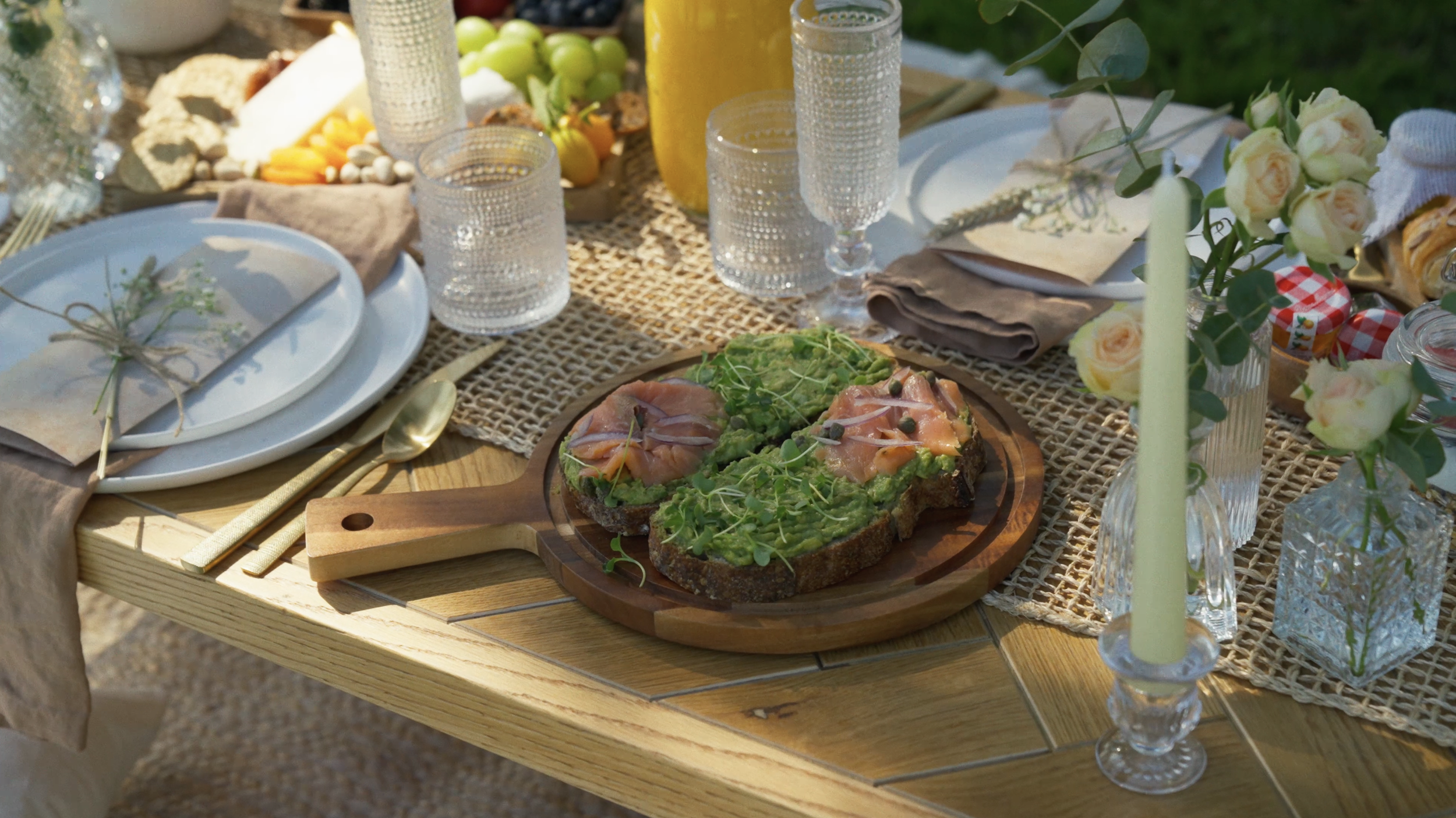 A rustic outdoor dining table set with a woven table runner, plates, glasses, a cutting board with avocado toast topped with microgreens and slices of prosciutto, and floral decorations including roses and greenery.