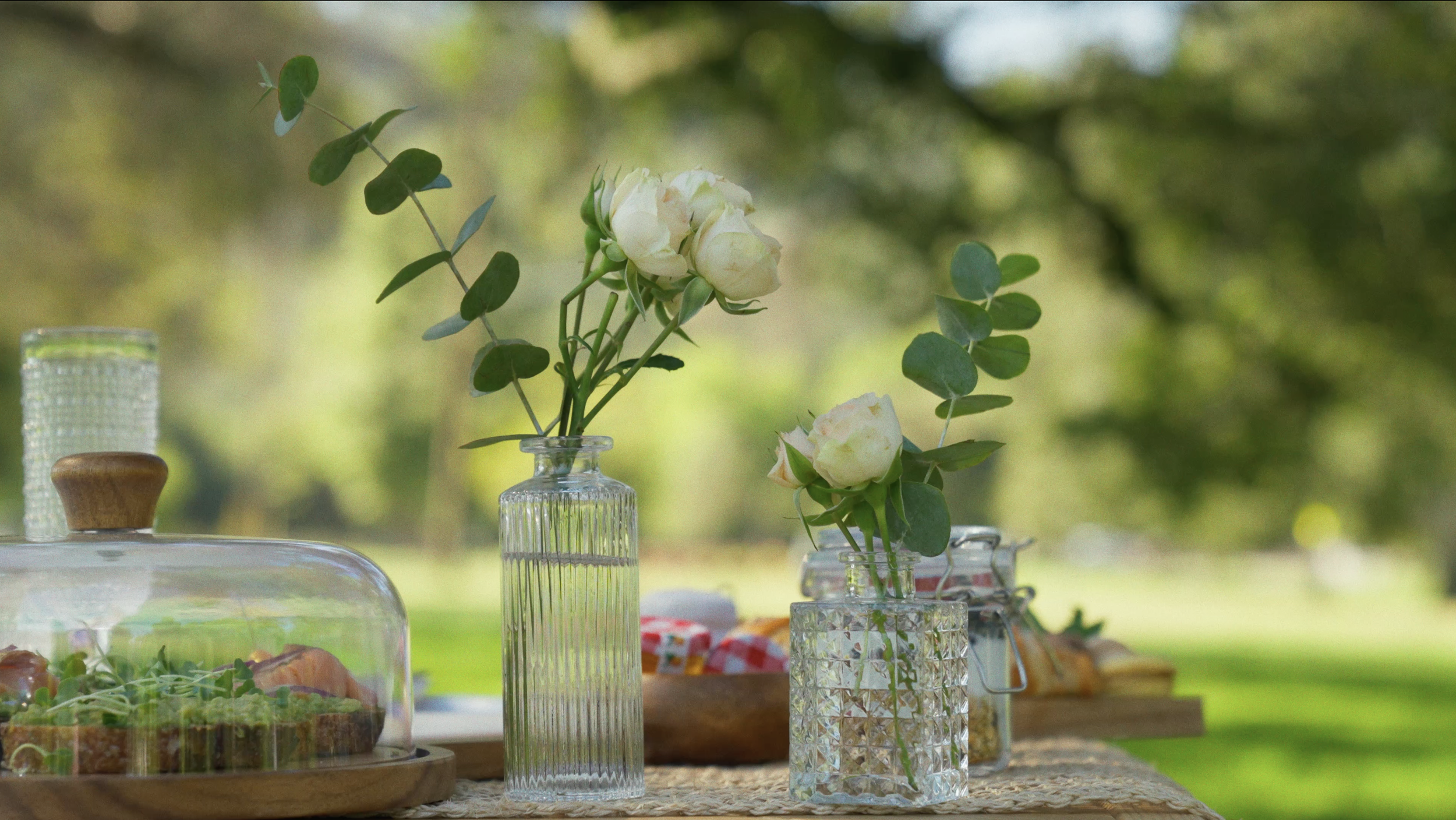 White roses in small glass vases on a table outdoors with greenery in the background.