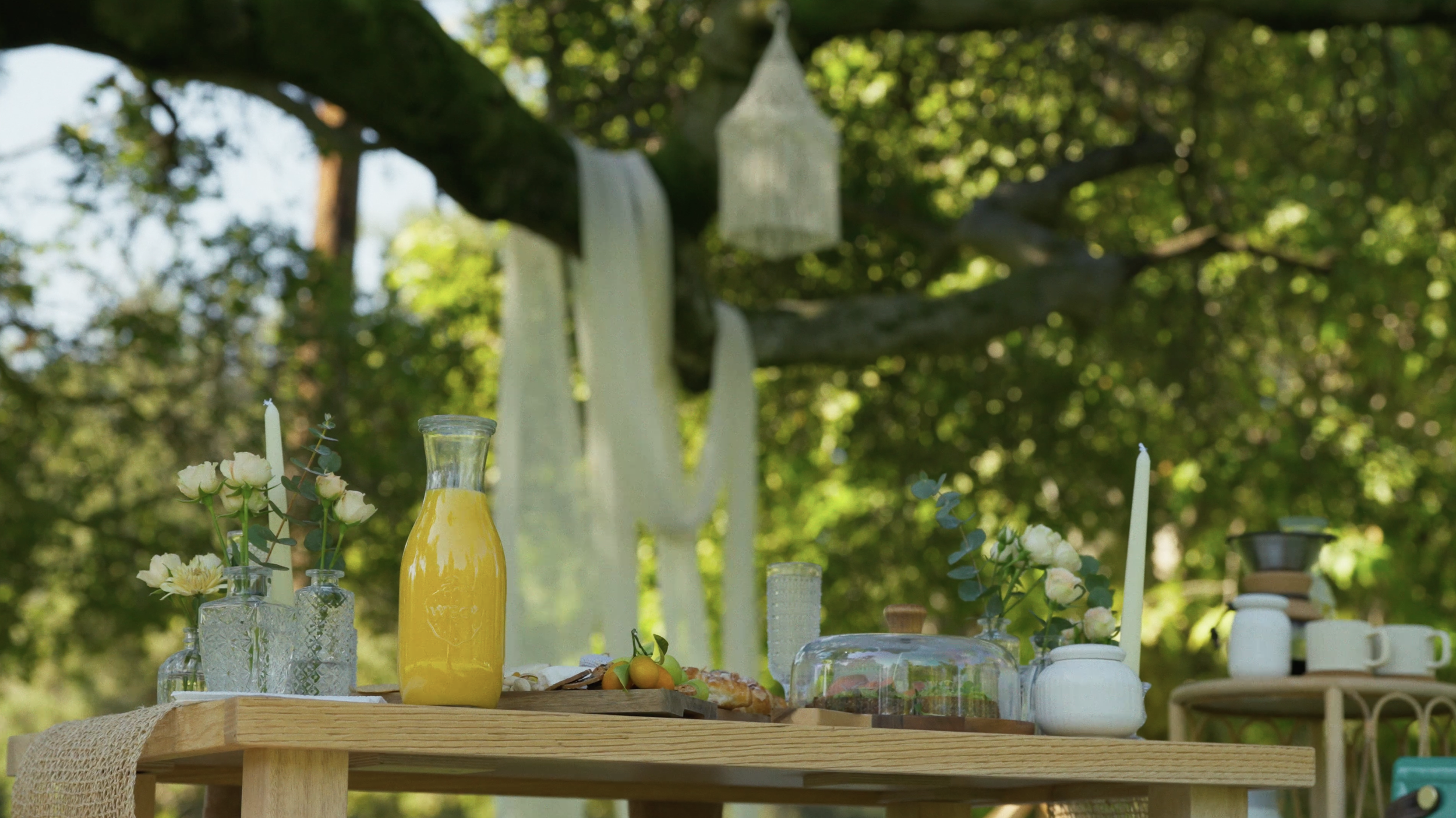 Outdoor table set with flowers, a yellow drink, and candles, under a tree with hanging lanterns.