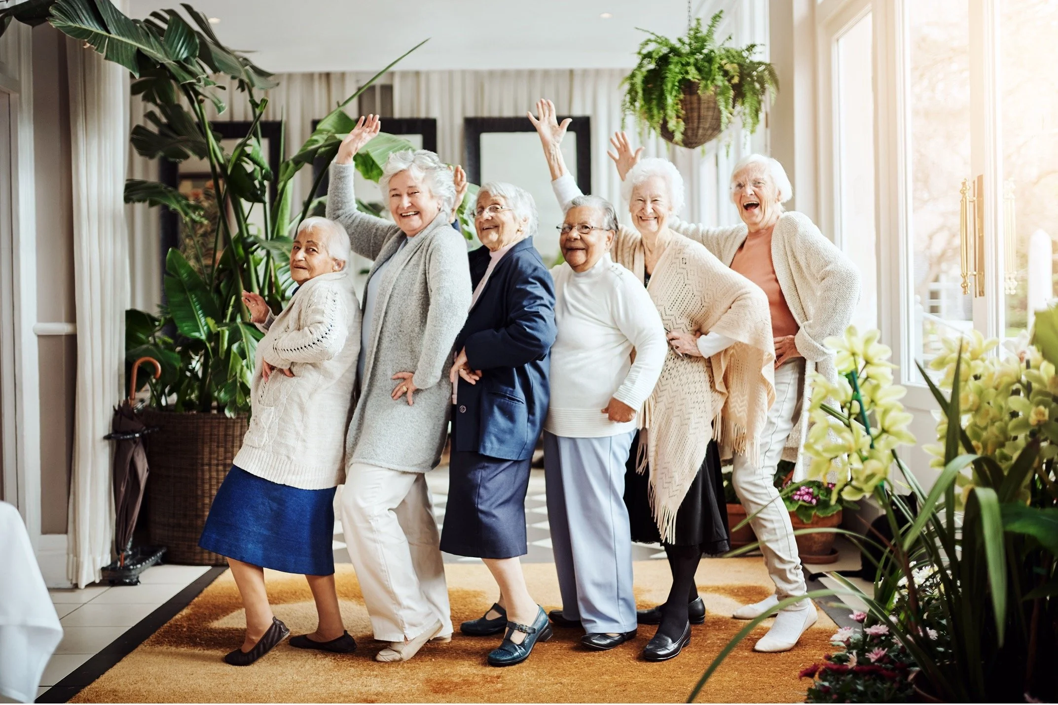 Group of elderly women happily posing and dancing together in a bright living room with large windows, potted plants, and floral decorations.