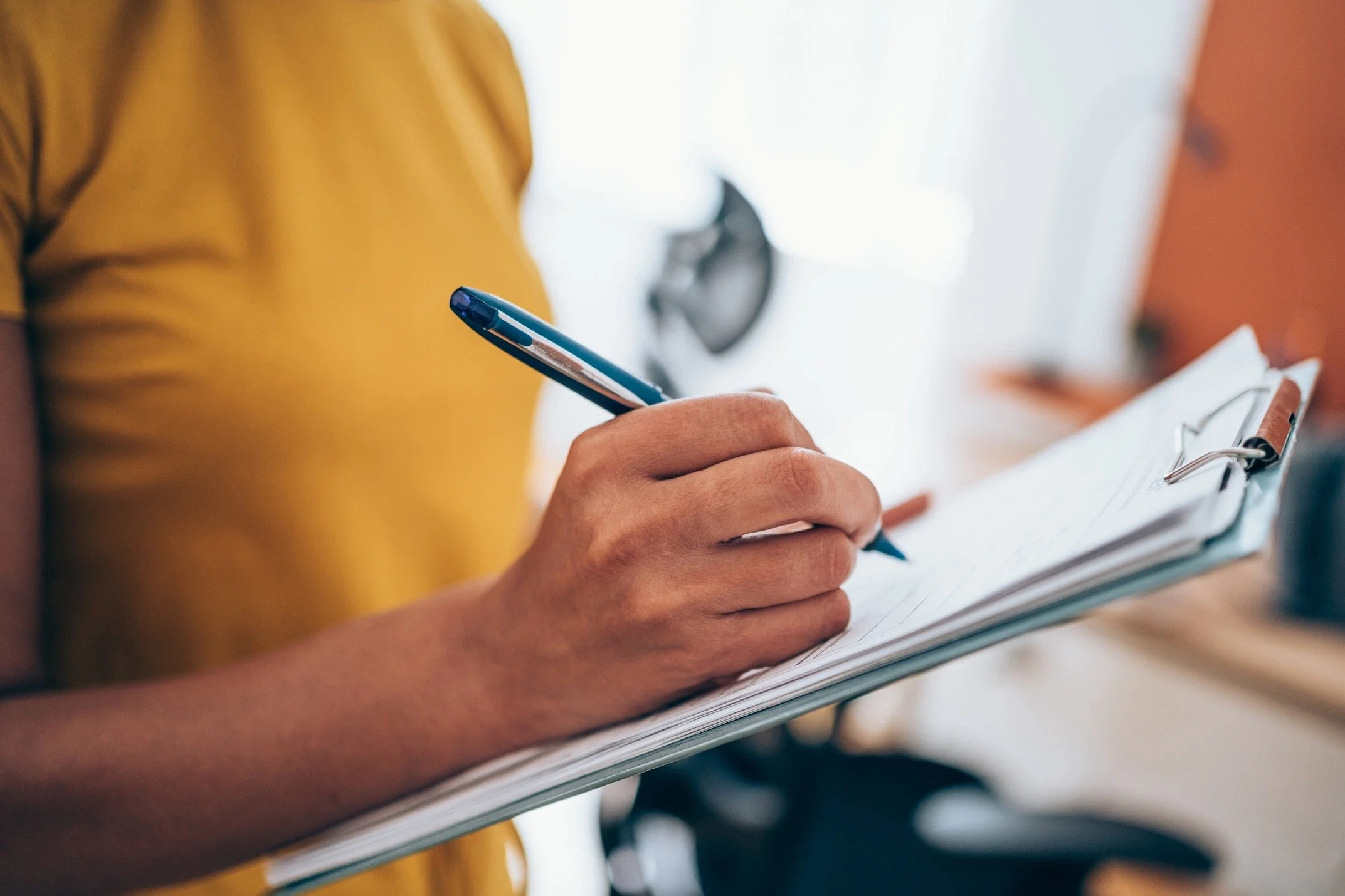 A person wearing a yellow top writing on a clipboard with a blue pen.