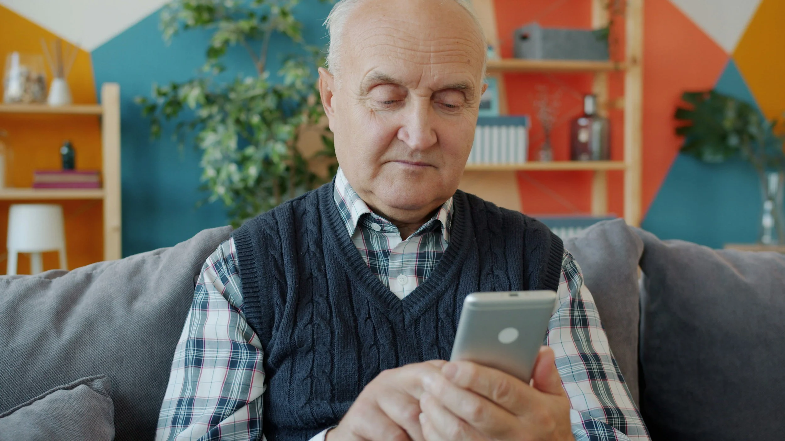 An elderly man with closed eyes, sitting on a gray couch, looking at his smartphone in a colorful room with bookshelves, plants, and wall decorations.