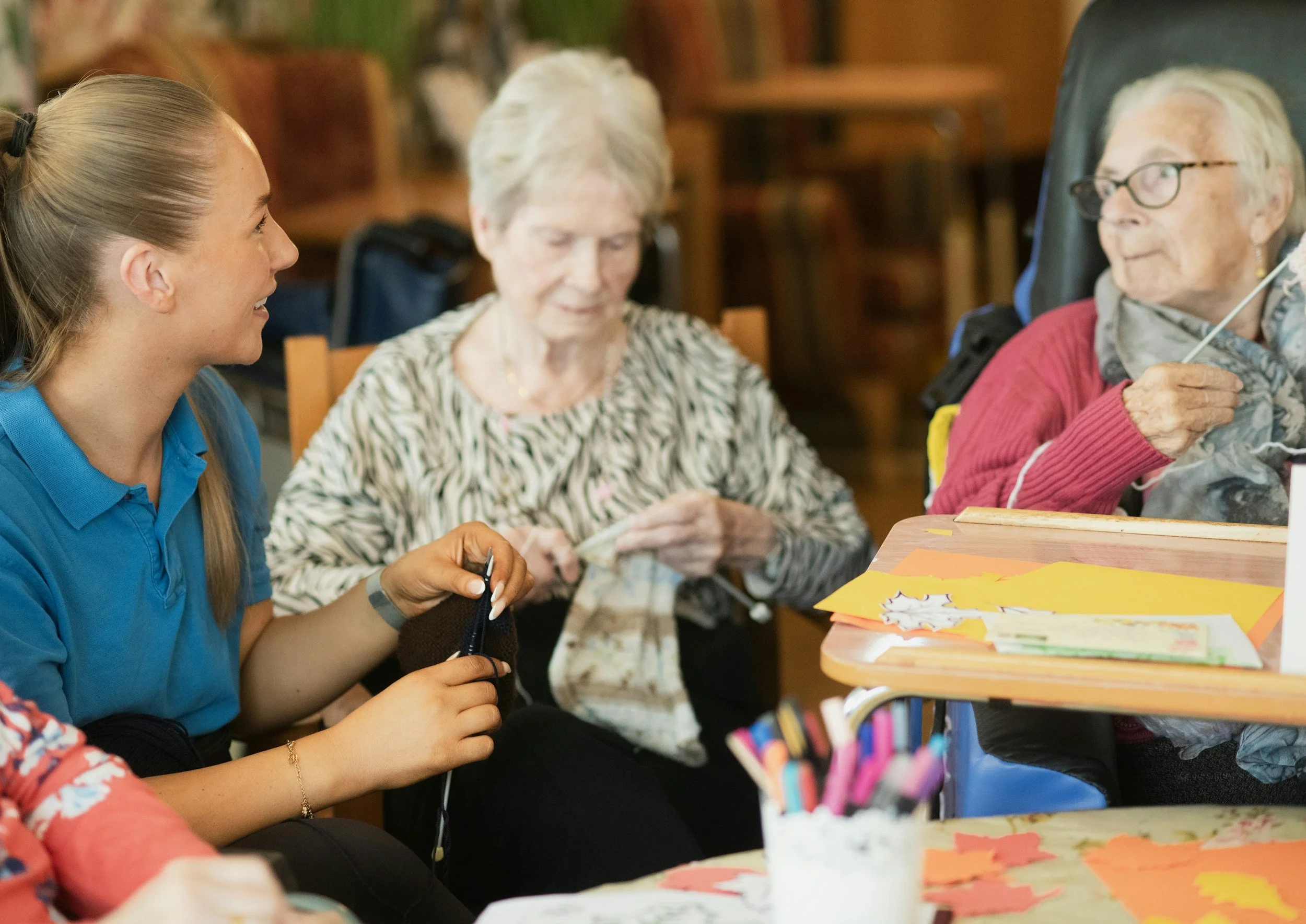 A young woman in a blue polo shirt assisting elderly women in a room with wooden chairs and tables. One elderly woman is holding a piece of cloth or fabric, and another is seated with a knitting project. There are craft supplies on the table, including colored paper, markers, and snacks.