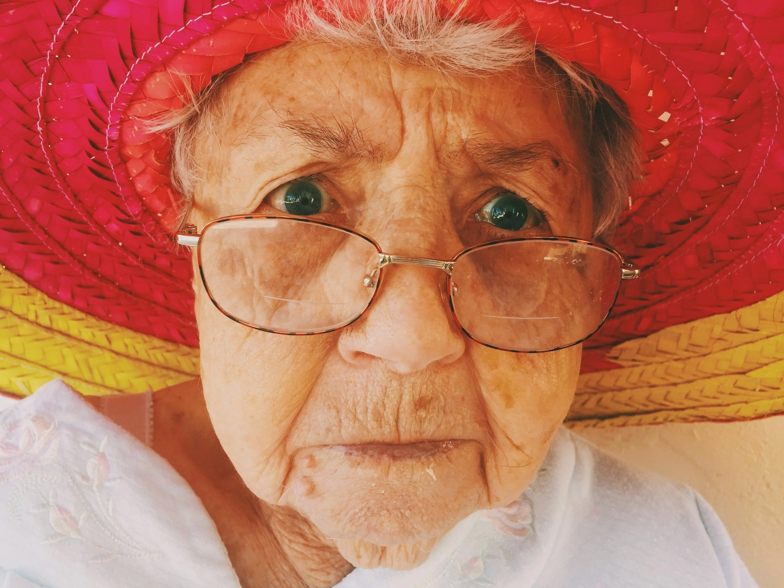 Close-up of an elderly woman wearing glasses and a colorful woven sombrero, with a serious facial expression.