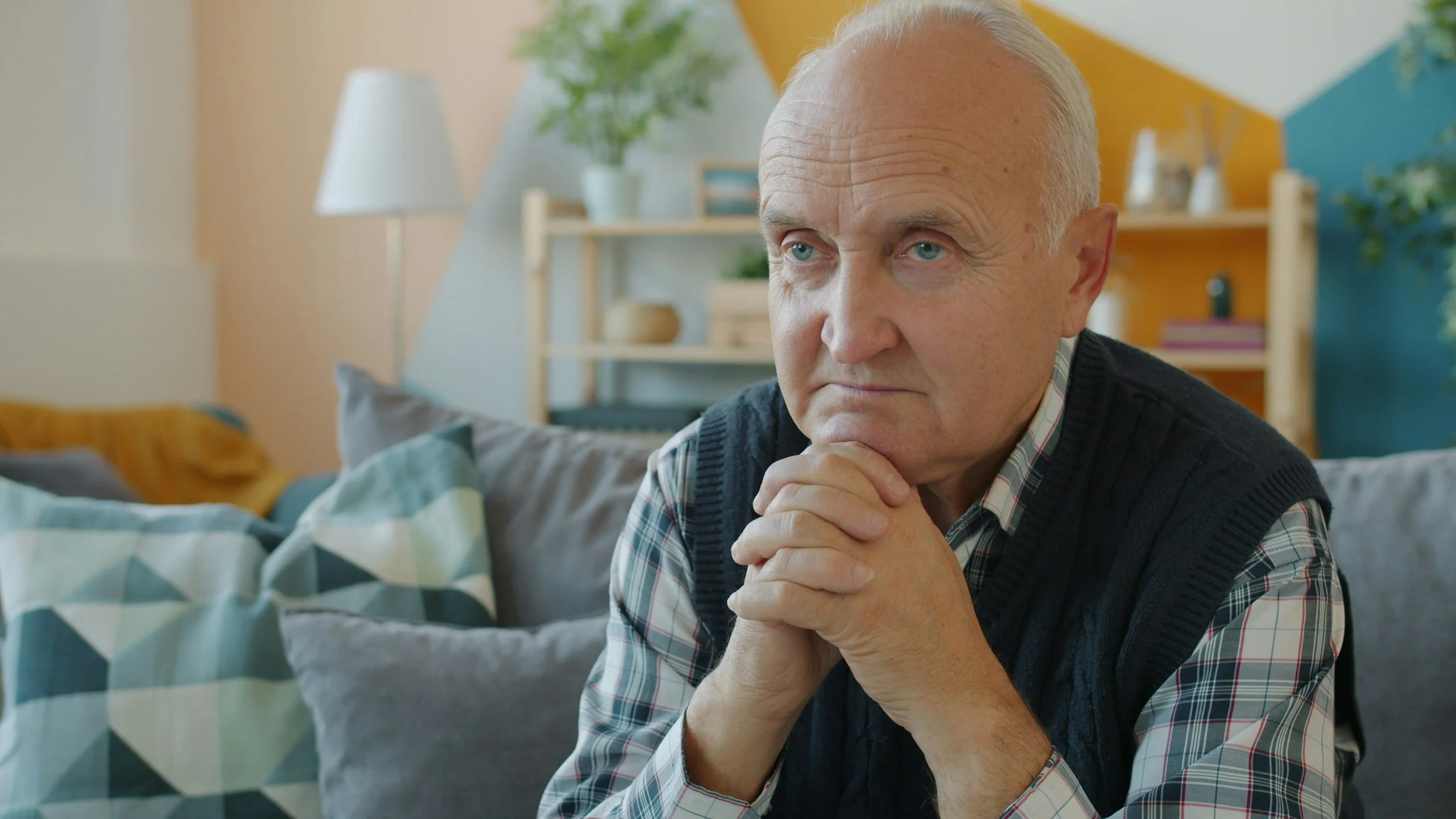 An elderly man with white hair and blue eyes sitting on a gray sofa in a modern, colorful living room. He is resting his chin on clasped hands, appearing thoughtful.