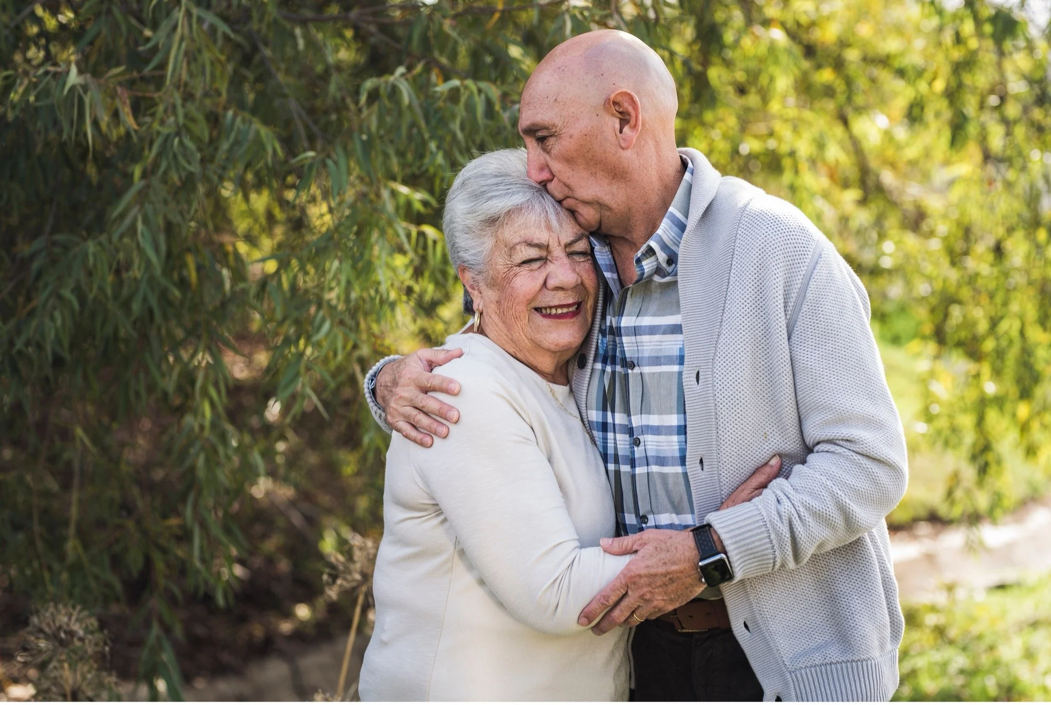 An elderly woman and a middle-aged man embracing outdoors, with the man kissing the woman's forehead. Both are smiling and appear to be enjoying a tender moment in a natural setting with trees and sunlight.