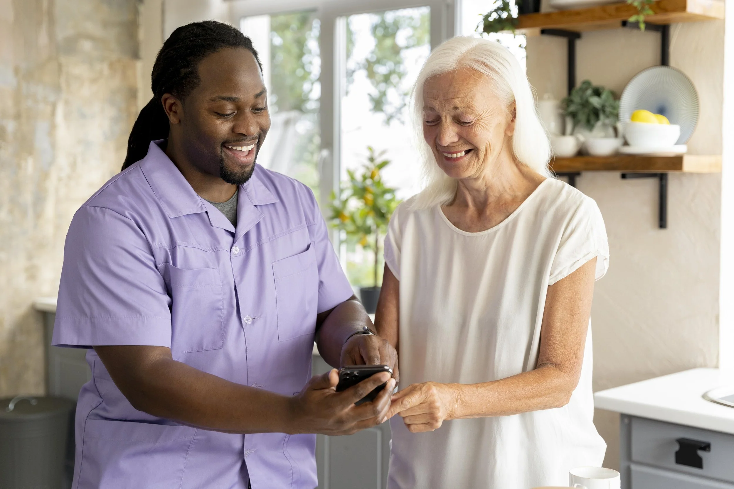 A young man in a purple shirt showing an older woman in a white dress something on his phone, both smiling in a bright kitchen.