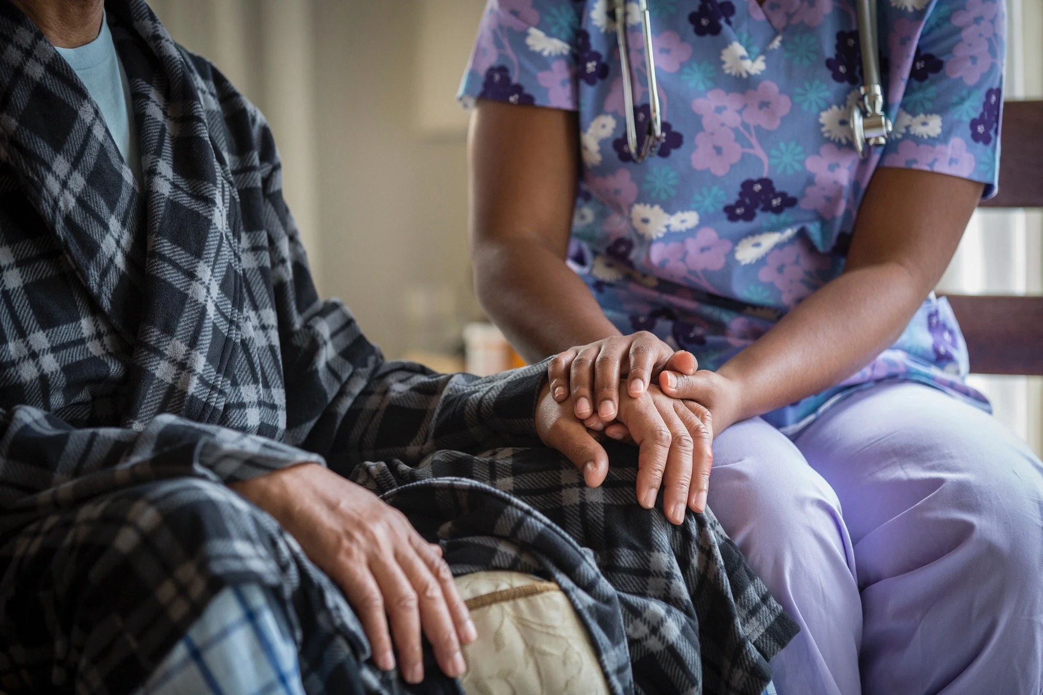 A nurse in scrubs gently holding the hand of a patient wearing a plaid shirt, sitting on a bench.