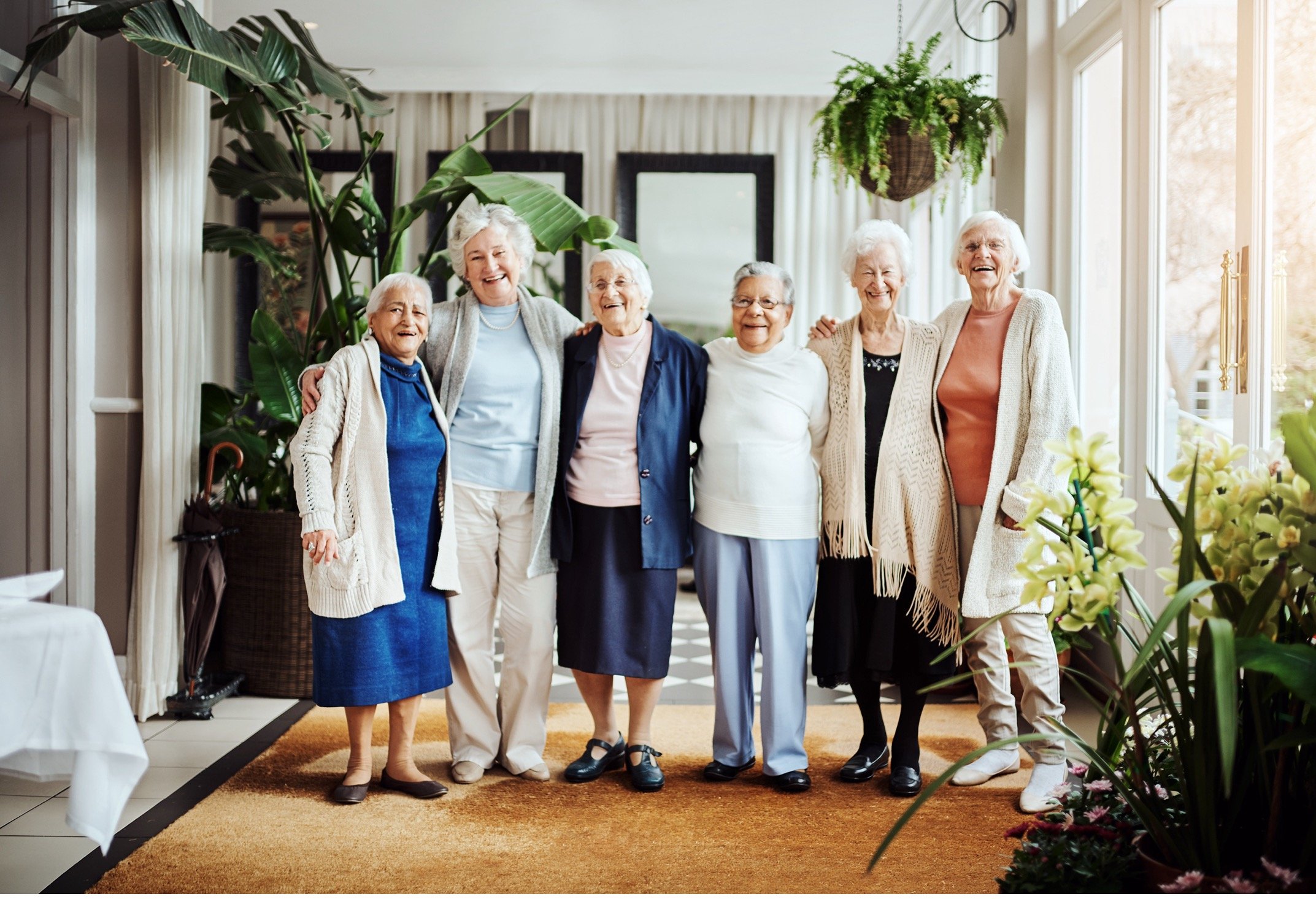 Seven elderly women standing together indoors, smiling and embracing each other, surrounded by green plants and sunlight.