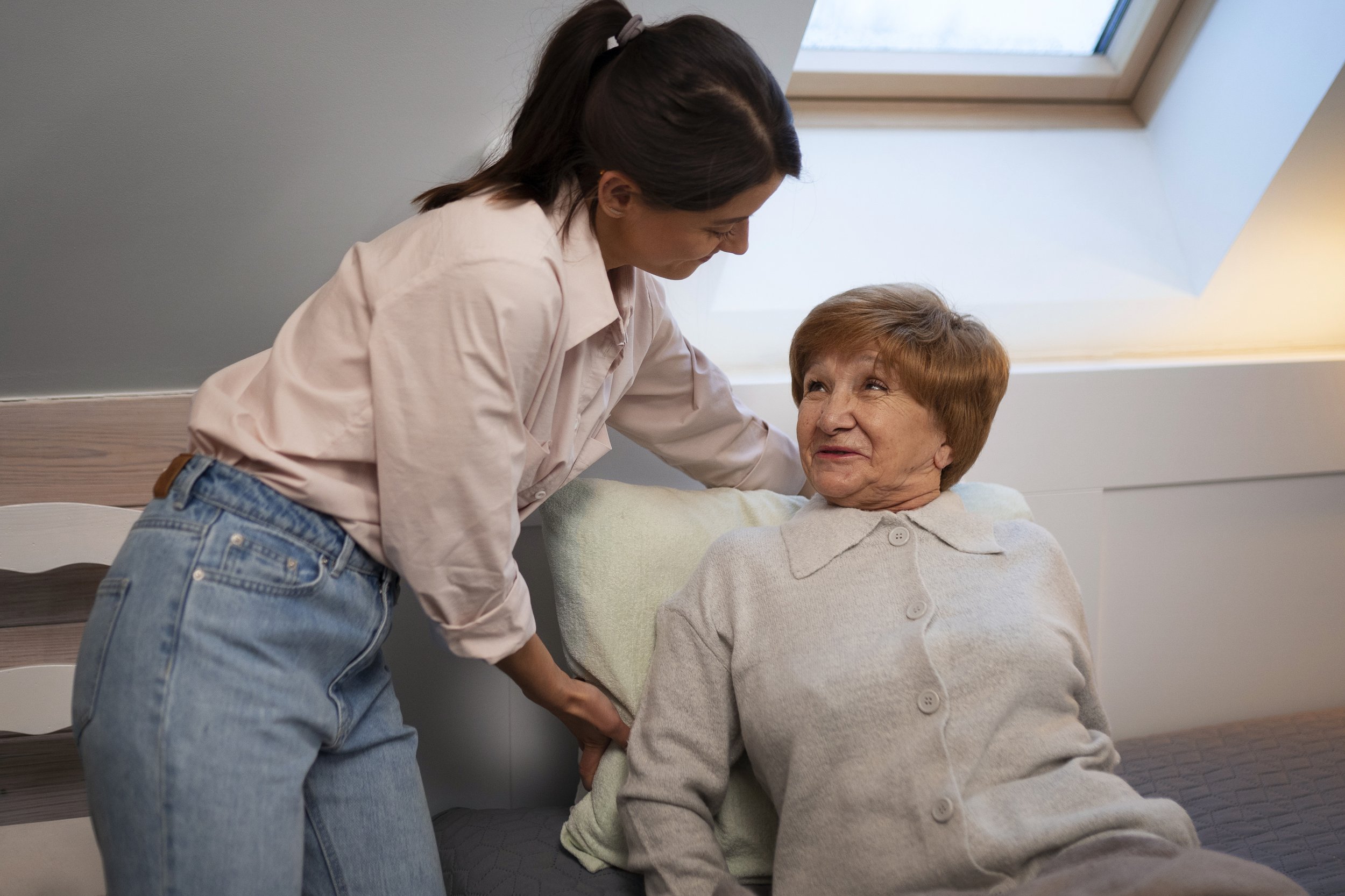 A young woman assists an elderly woman in bed, holding her waist gently, in a well-lit room with a window overhead.
