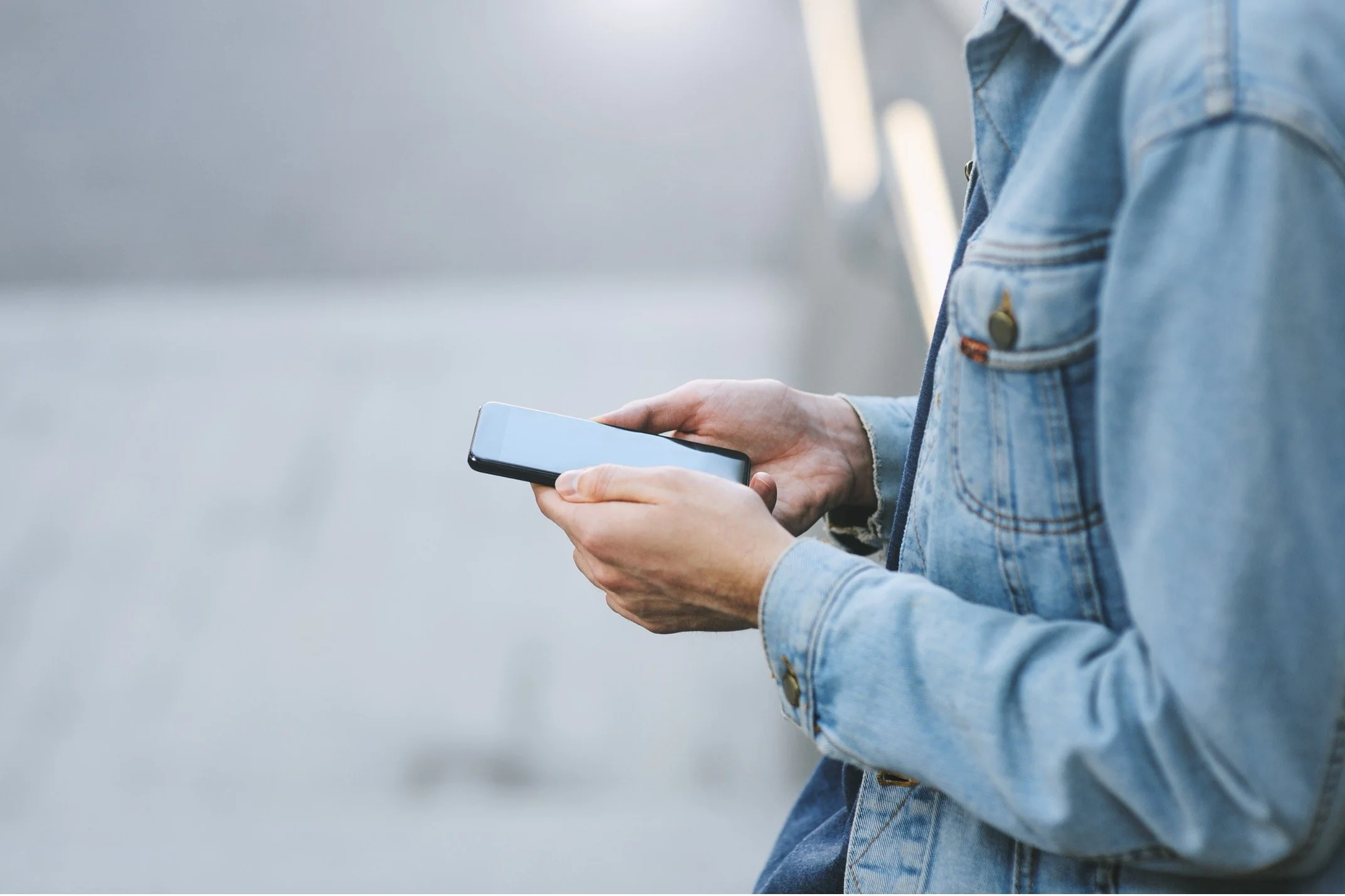 Person in a denim jacket holding and looking at a smartphone outdoors.