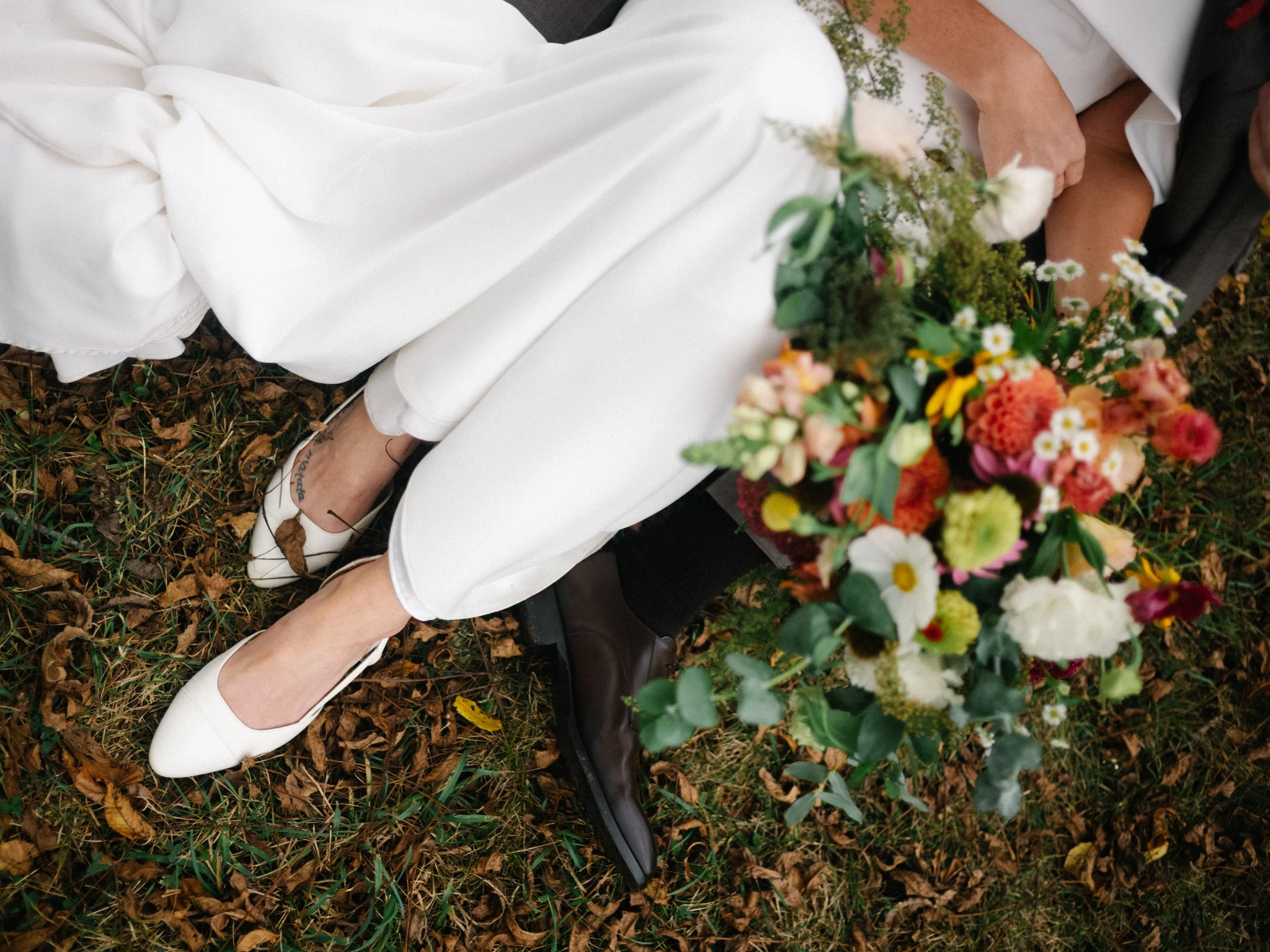 Couple sitting on a bench in a park, holding a large colorful bouquet of flowers, with fallen leaves on the ground, dressed in light-colored clothes.