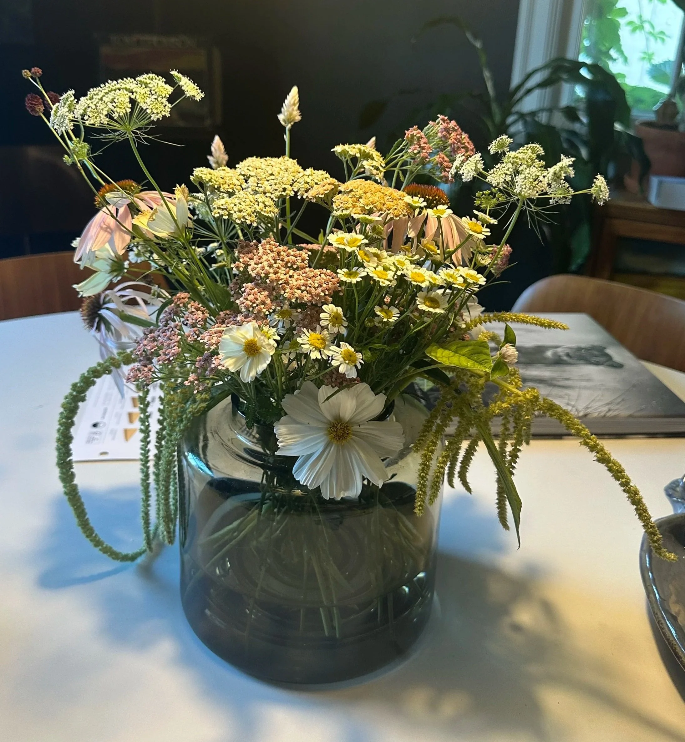 A bouquet of mixed flowers in a dark glass vase on a white table, with a window and houseplants in the background.