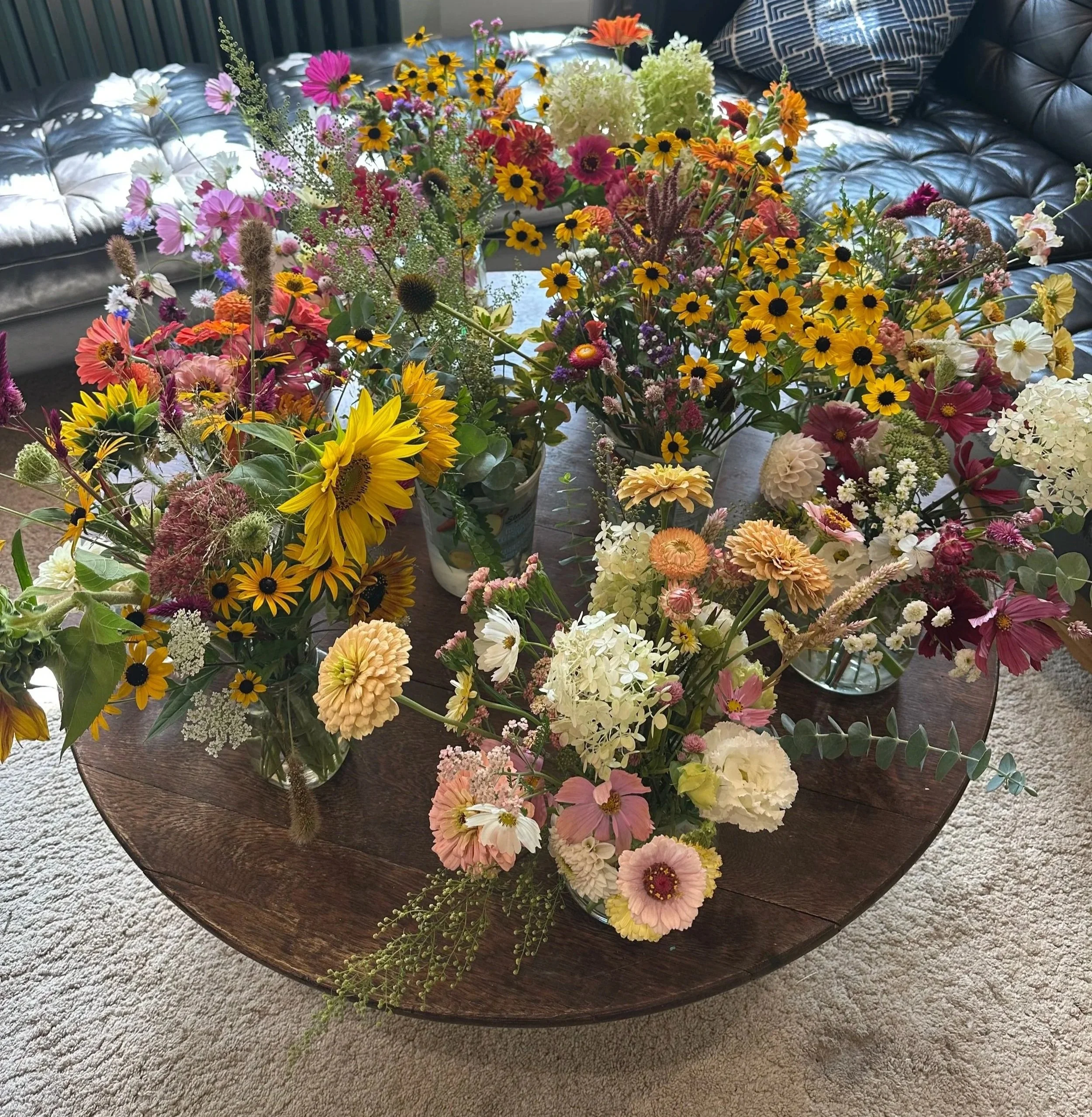 Multiple colorful flower arrangements in glass vases on a wooden table.