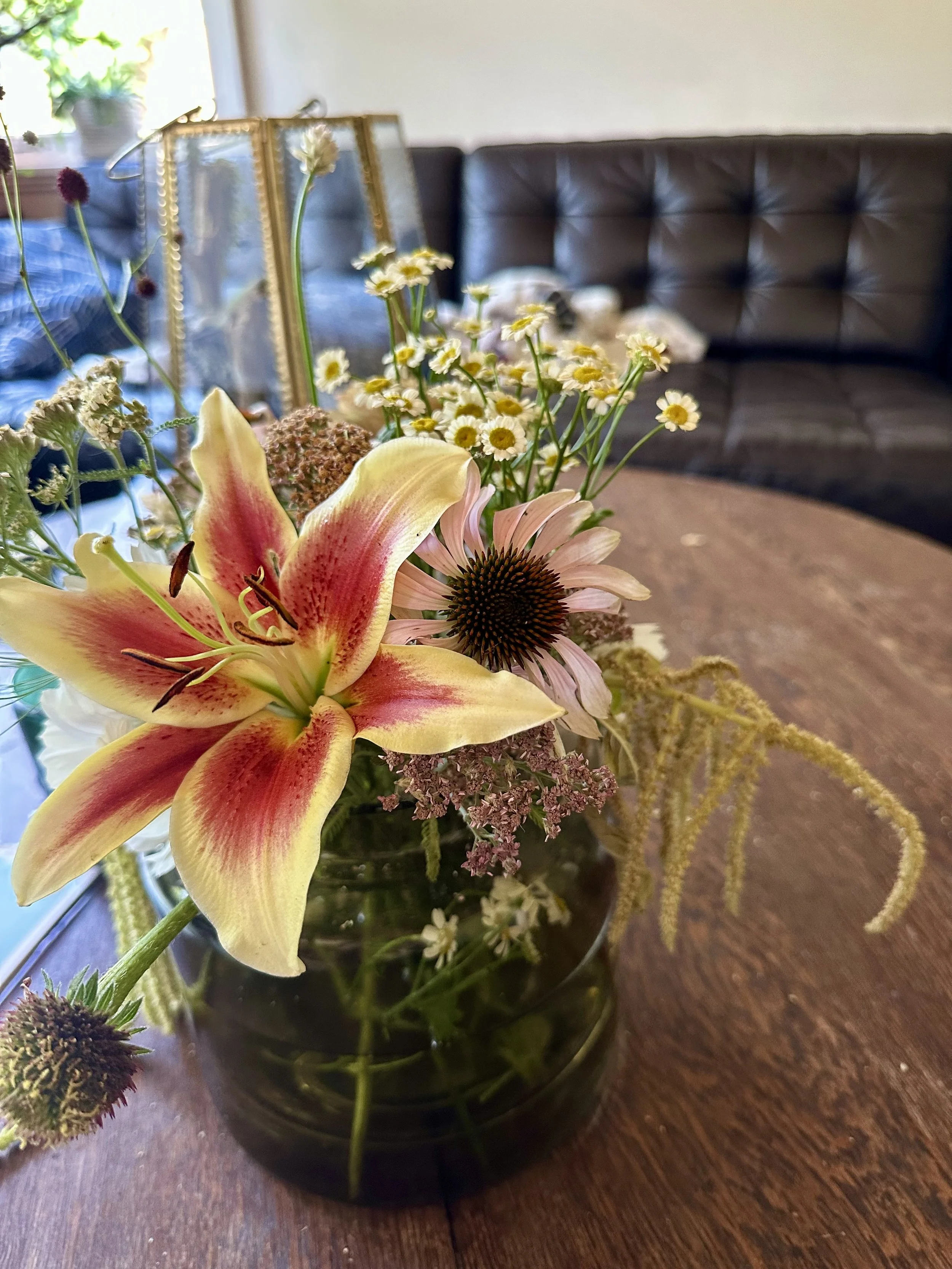 A bouquet of assorted flowers in a glass vase on a wooden table, with a blurred background of a brown tufted sofa and a window with plant.