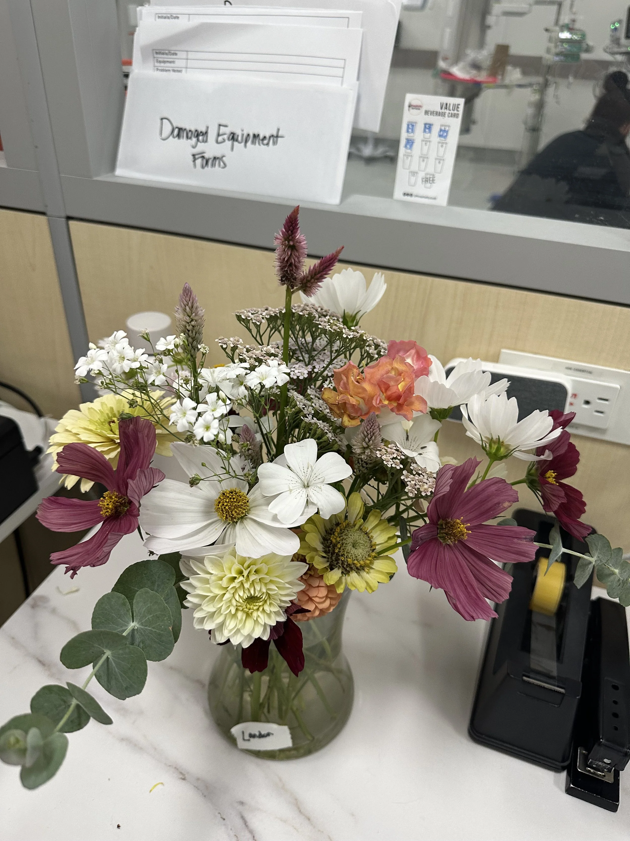 Colorful flower bouquet in a glass vase on a white countertop in a workspace, with signage and office equipment in the background.
