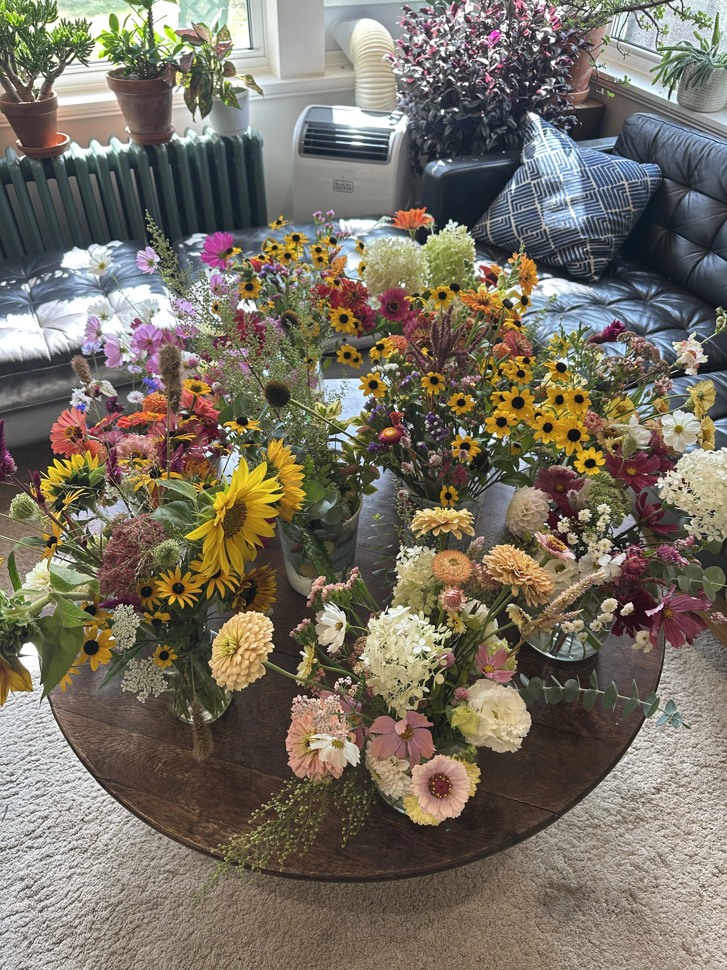Multiple colorful flower arrangements on a wooden table in a bright living room with plants on the windowsill and a leather couch with patterned pillows.